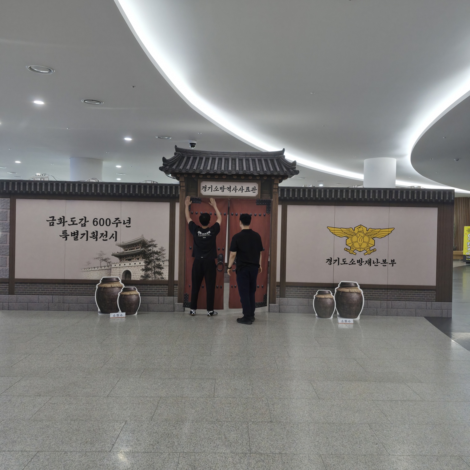 한옥 전통 건축 모양의 포토존 구조물. 목재와 어두운 색의 프레임으로 이루어진 한옥 형태이며, 상단에 기와지붕 장식, 중앙에 붉은색 문짝이 있고, 양쪽 패널에 흰색 배경의 인쇄 그래픽과 전통 항아리 스탠드 설치됨