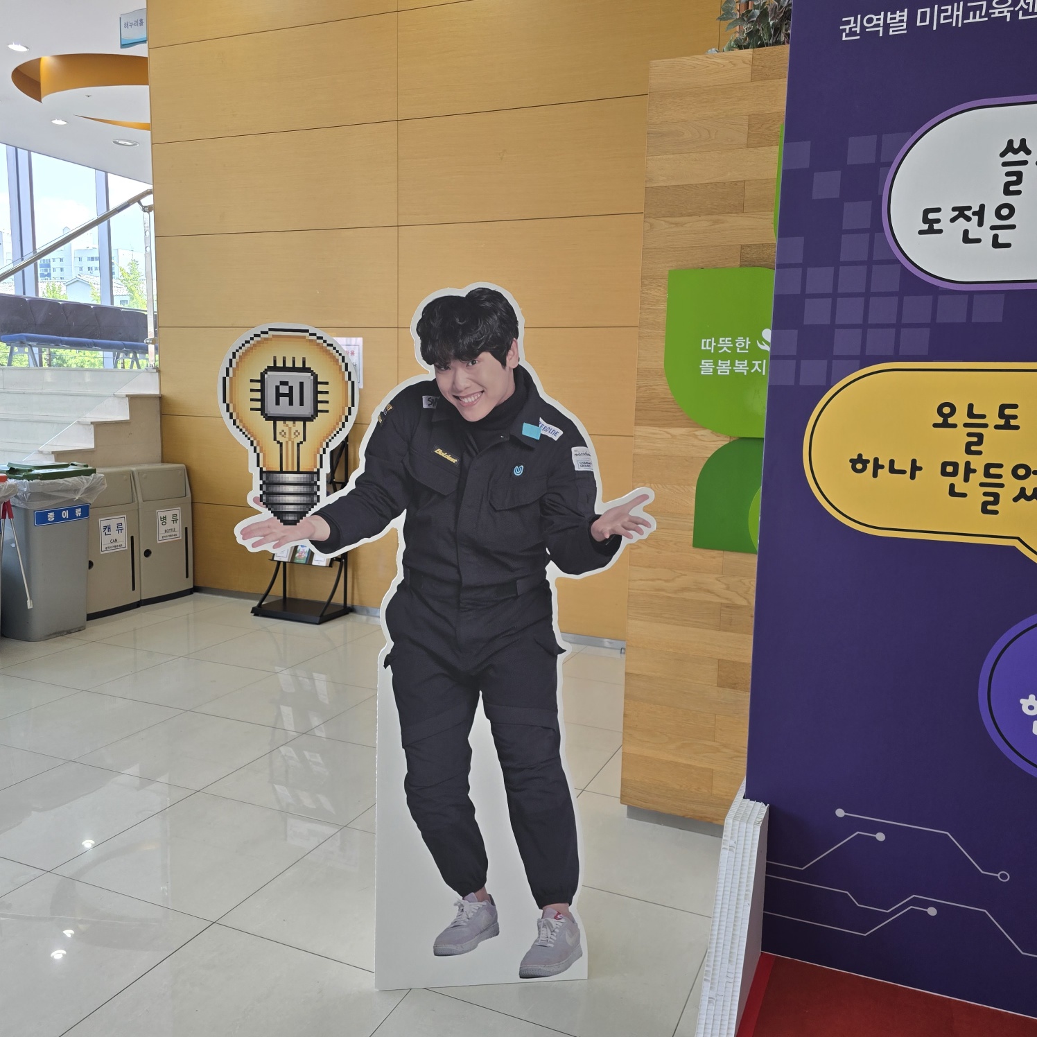 검은 유니폼을 입은 사람 옆에 AI 전구 모양 스카시가 세워져 있고, 뒤쪽에 말풍선 스티커와 보라색 판넬이 보임. 경비실과 나무 벽 앞에 배치됨.