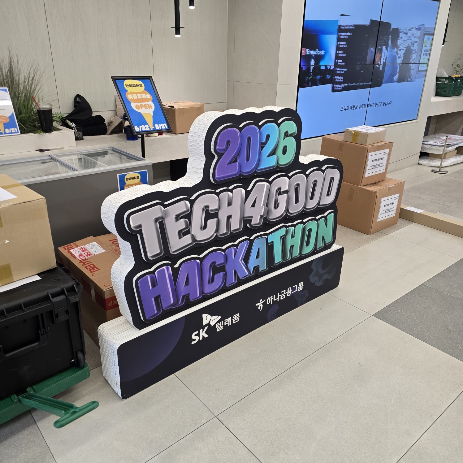 앞면이 흰색 메시 재질인 대형 입체 조형물. '2026 TECH4GOOD HACKATHON' 텍스트가 검은색 테두리와 함께 그래디언트 색상으로 표현되었으며, 하단부에 검정 배경에 하나금융그룹과 SK 텔레콤 로고가 인쇄되어 있음
