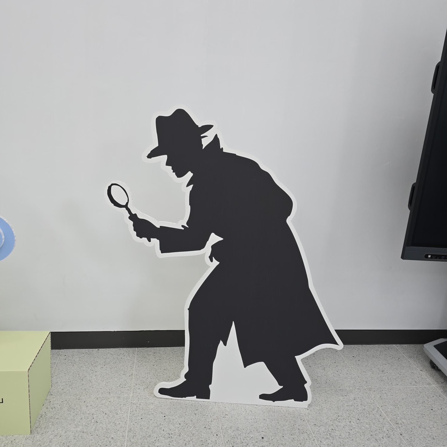 검은색 실루엣으로 제작된 탐정 캐릭터 스카시. 모자와 코트를 입고 돋보기를 든 탐정 인물이 몸을 숙인 자세. 흰색 테두리로 외곽선이 처리되어 벽에서 분리되어 보임. 자립형으로 바닥에 설치됨