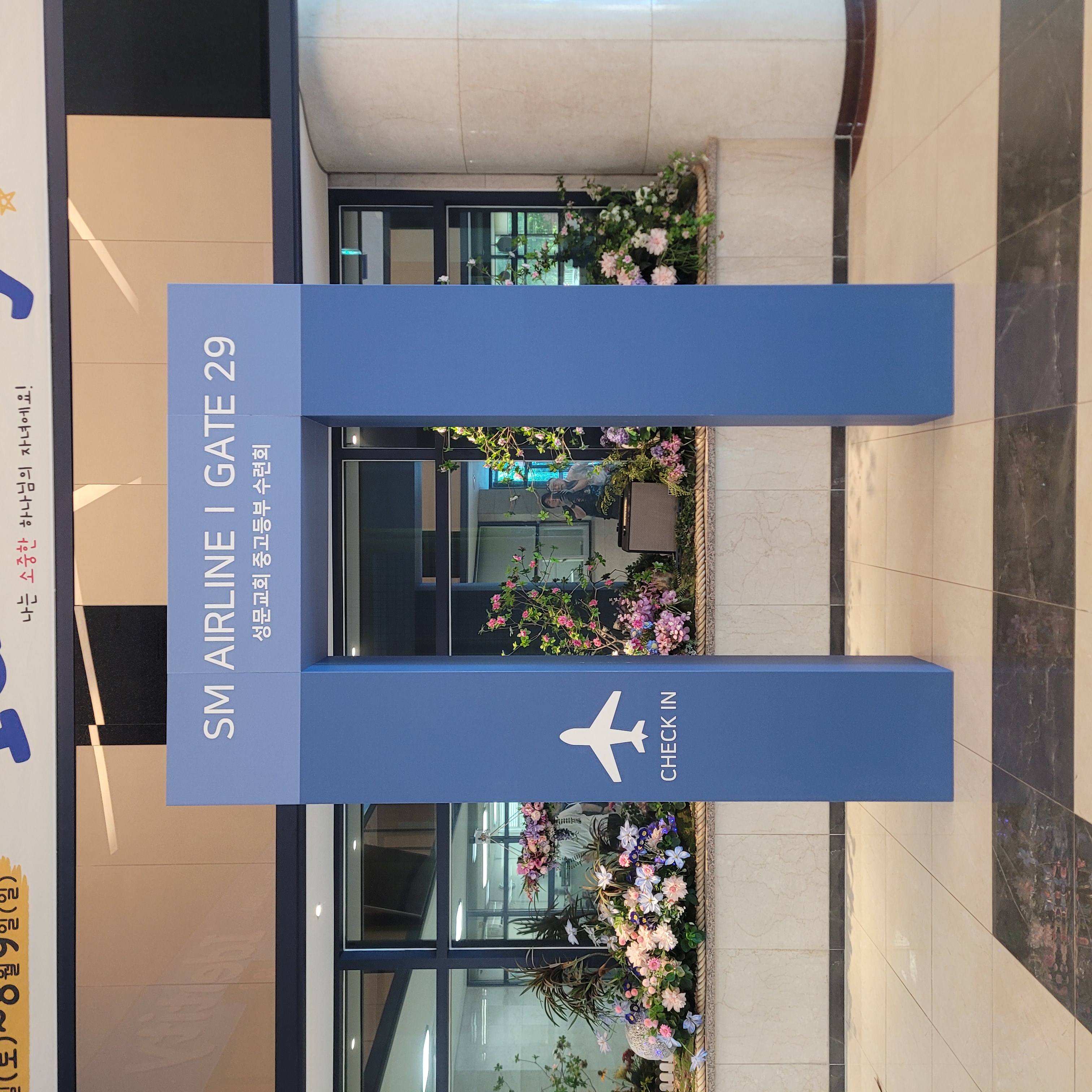 파란색 사각 기둥 2개로 구성된 입구 게이트. 상단에 'SM AIRLINE | GATE 29' 'CHECK IN' 문구와 비행기 아이콘이 인쇄됨. 내부에 꽃 장식과 체크인 공간이 보임.