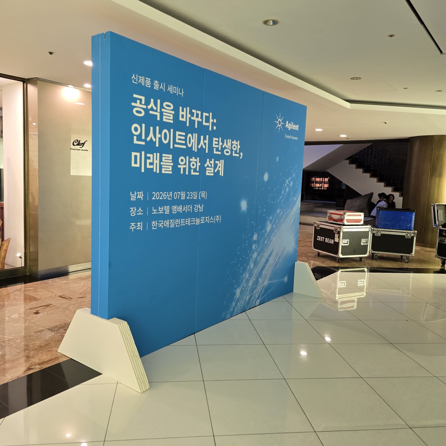 측면에서 본 같은 패널. 파란색 인쇄면에 세미나 제목·일정 표기, 하단 허니콤 지지대 형태가 뚜렷이 보임. 실내 로비 바닥에 자립형으로 설치되어 있고 뒤로 계단·인테리어가 보임.