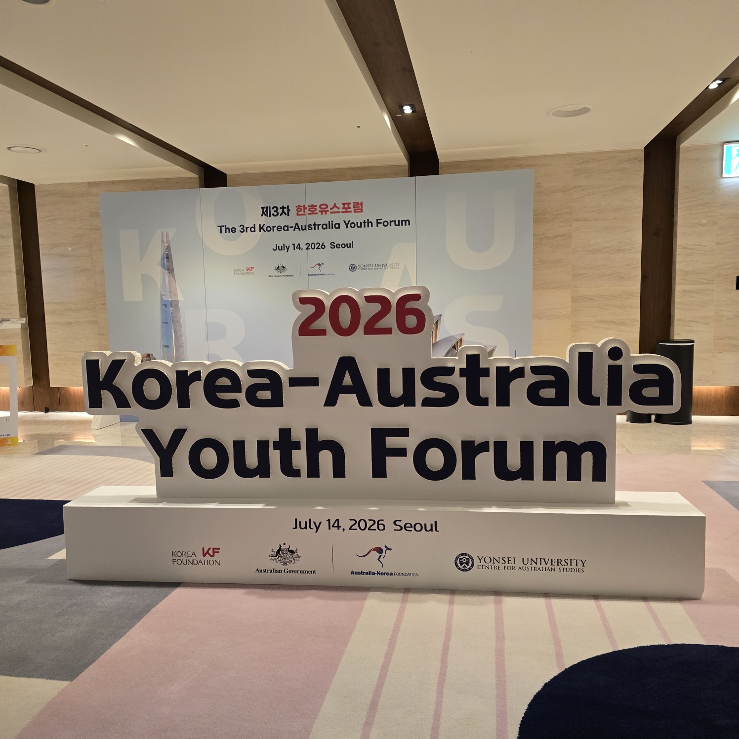 흰색 아크릴 받침대 위에 진한 파란색 입체 글씨로 '2026 Korea-Australia Youth Forum' 텍스트가 표현되어 있으며, 하단에 'July 14, 2026 Seoul'과 각 기관 로고가 인쇄된 받침판 구조