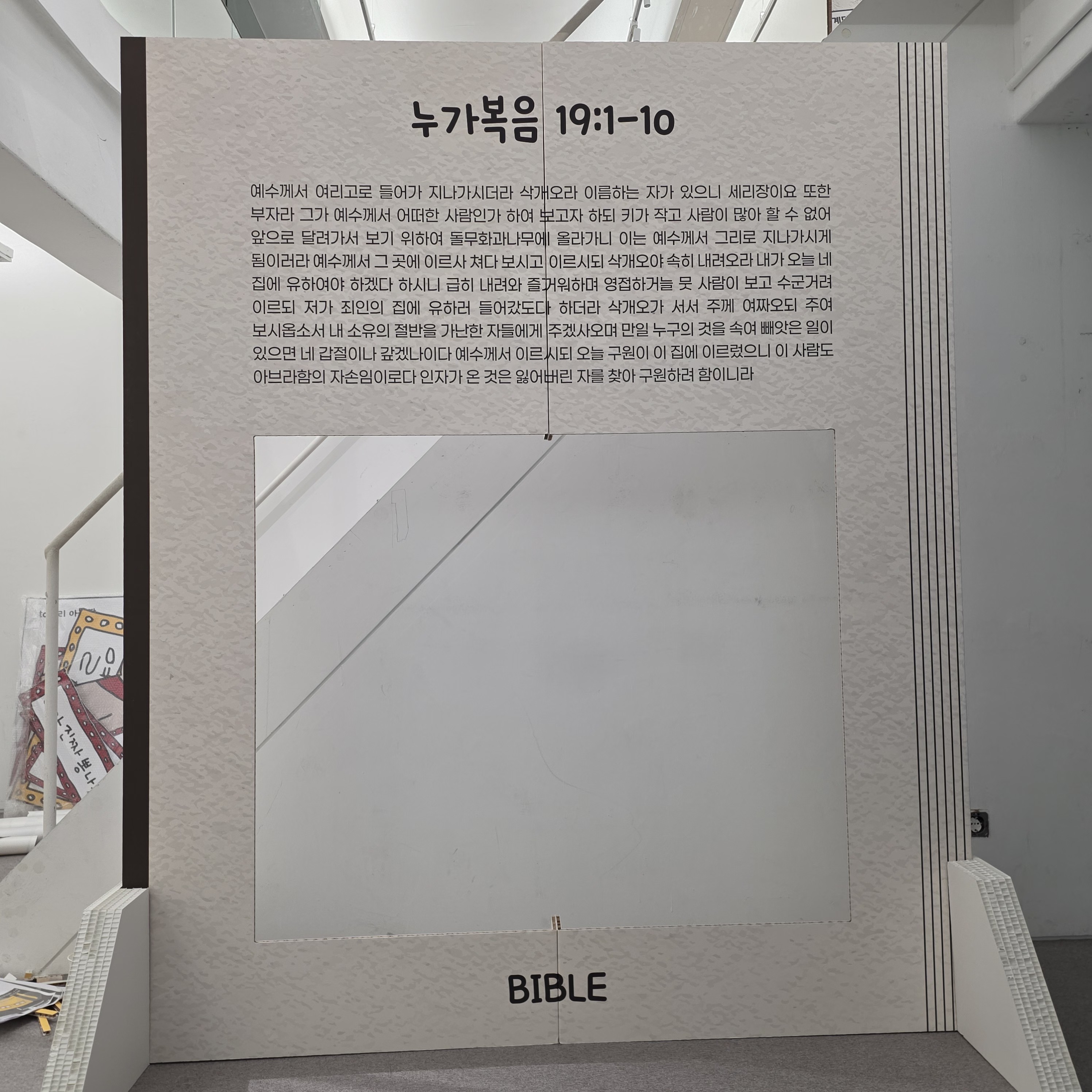 책 형태의 대형 포토존. 회색 표지에 '누가복음 19:1-10' 제목과 본문, 중앙에 사각 창문, 하단에 'BIBLE' 문구. 입체적인 책 페이지 형상.