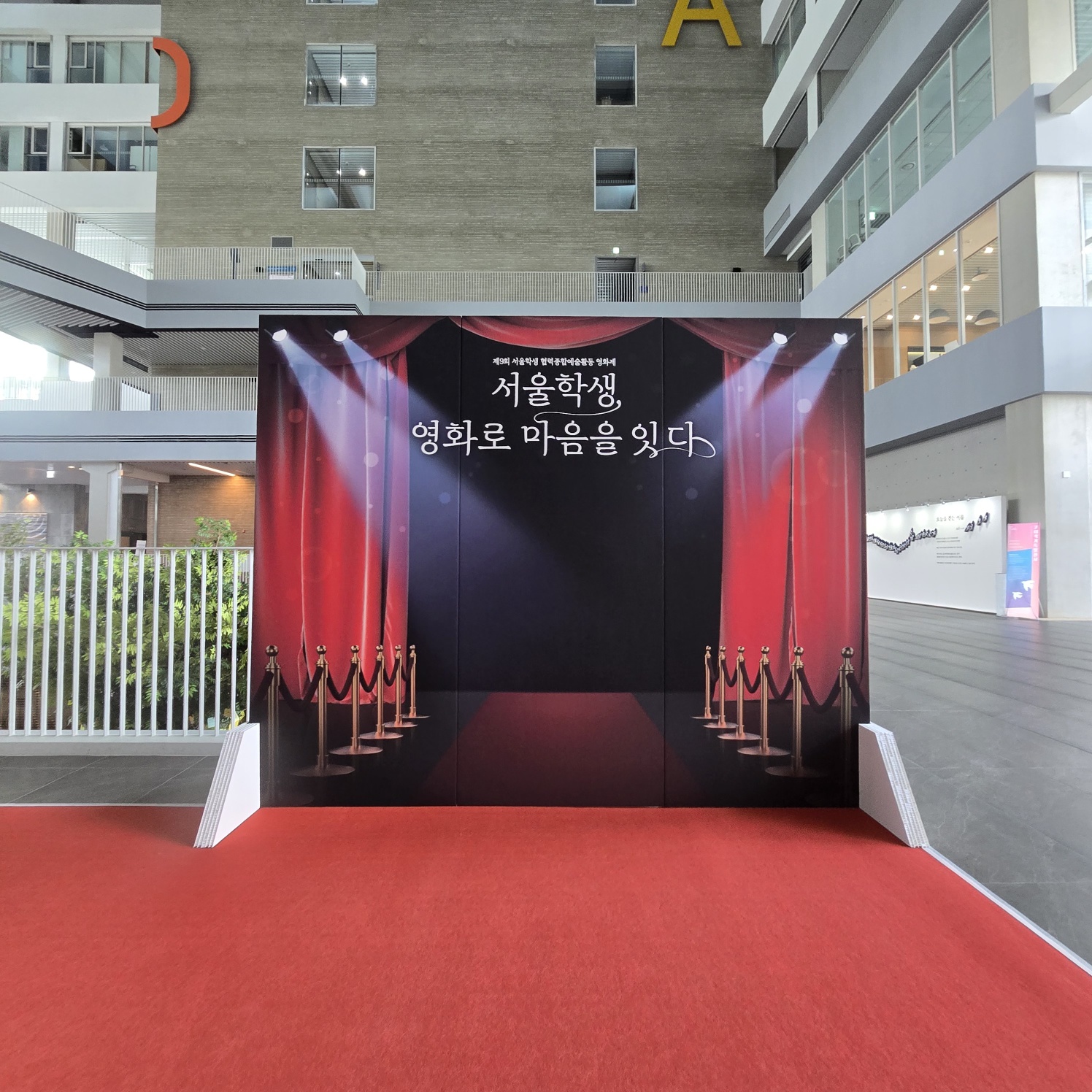빨간 무대 커튼과 스포트라이트가 인쇄된 검은색 배경 패널. 중앙에 '서울학생, 영화로 마음을 잇다' 문구가 흰색으로 새겨져 있고, 하단에 빨간 카펫과 벨벳 로프 기둥들이 그려져 있음. 양쪽에 흰색 삼각 받침대가 보임.