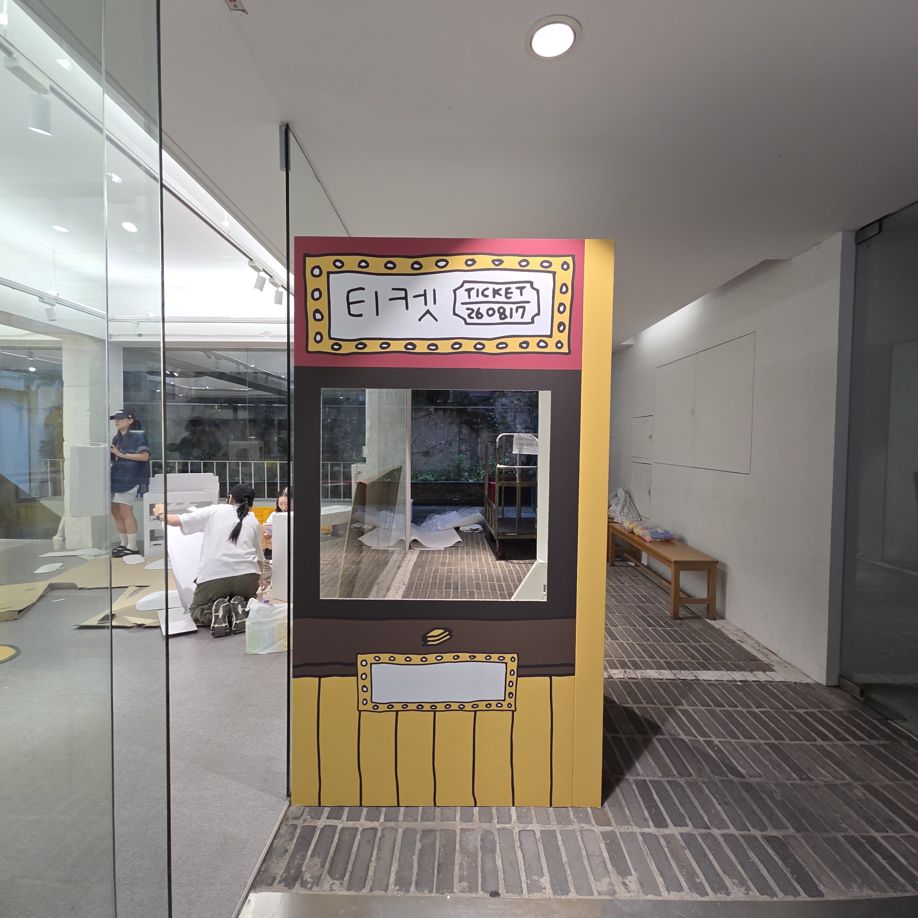 노란색 바탕에 '티켓 260817' 문구와 장식 테두리가 인쇄된 포토존. 중앙에 사각 창문이 뚫려 있고, 하단은 나무판자 질감으로 마감. 입체적으로 자립하는 구조.