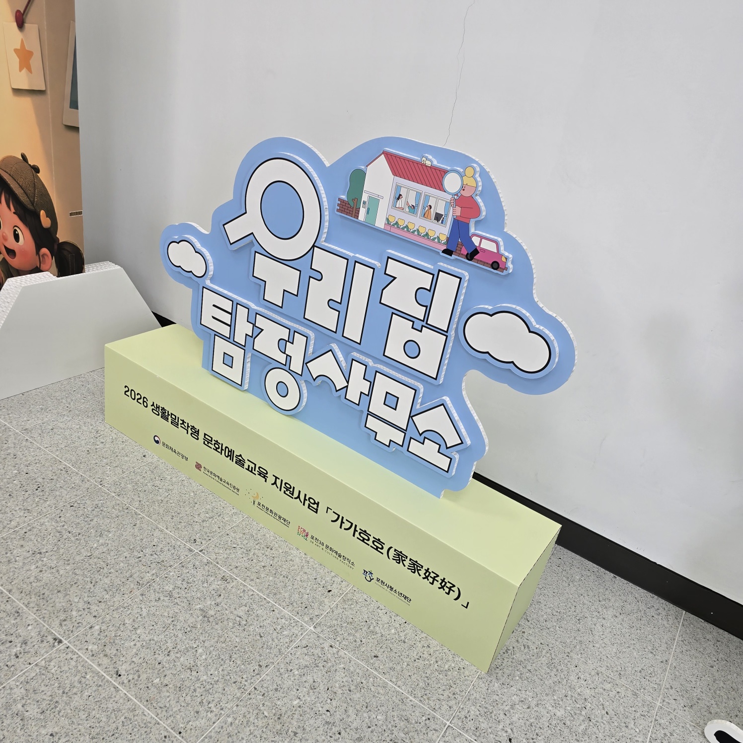 측면 각도에서 본 하늘색 구름형 스카시. 입체 글씨와 일러스트의 두께감이 보이며, 연두색 박스 받침대 위에 자립 설치됨. 허니콤보드 구조로 제작되어 경량이면서 안정적으로 서 있음