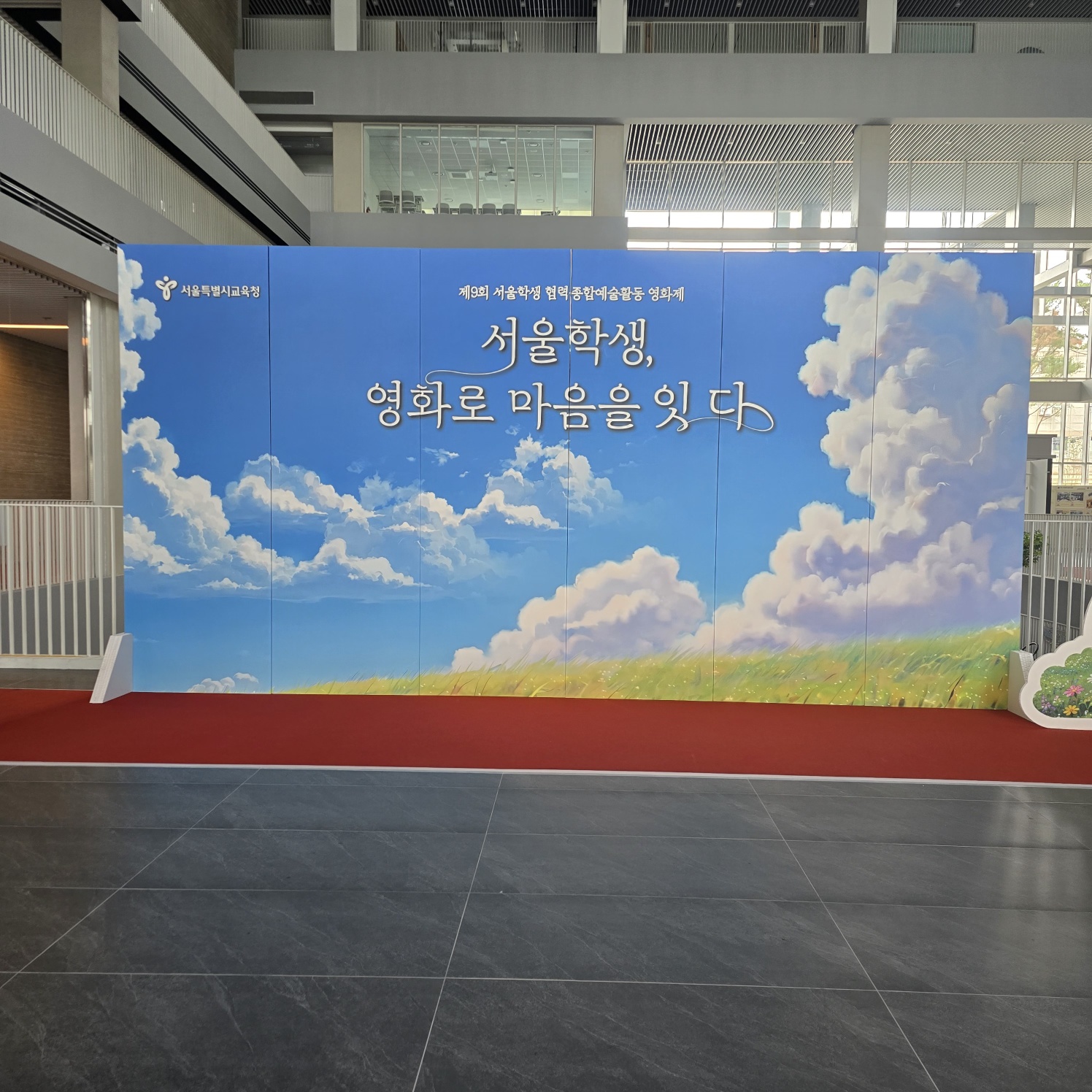 파란 하늘과 구름이 인쇄된 대형 배경 패널에 '서울학생, 영화로 마음을 잇다' 문구가 흰색으로 새겨져 있고, 하단에는 초록 잔디밭과 흰 꽃이 그려진 평평한 포토존 배경. 패널 앞 바닥에 빨간 카펫이 깔려 있고, 오른쪽에 꽃무늬 장식 소품이 보임.