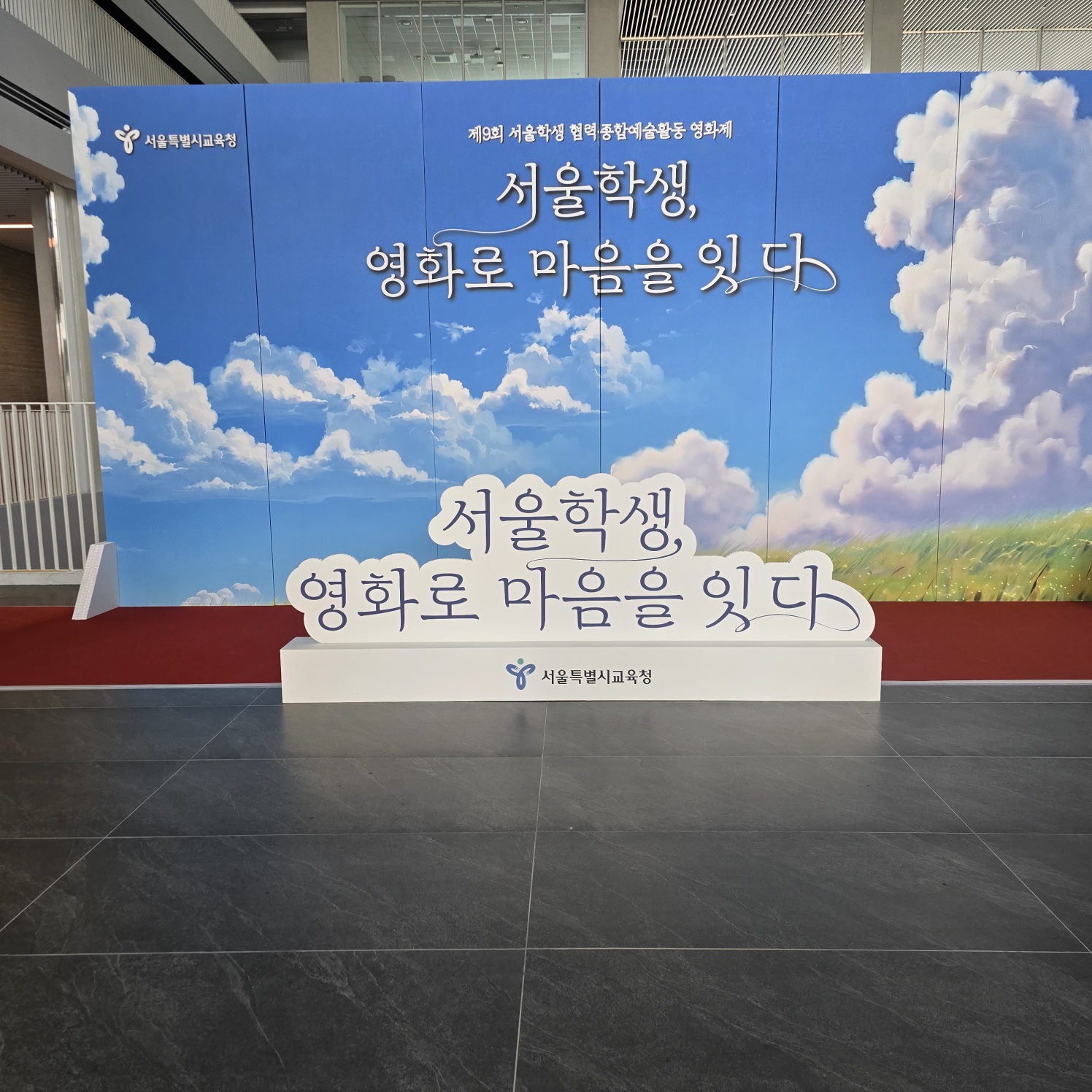 파란 하늘 배경 패널 앞 중앙에 구름 모양 흰색 입체 간판이 서 있고, '서울학생, 영화로 마음을 잇다' 문구와 '서울특별시교육청' 로고가 인쇄됨. 바닥에 빨간 카펫, 좌우에 흰색 삼각 받침대가 보임.