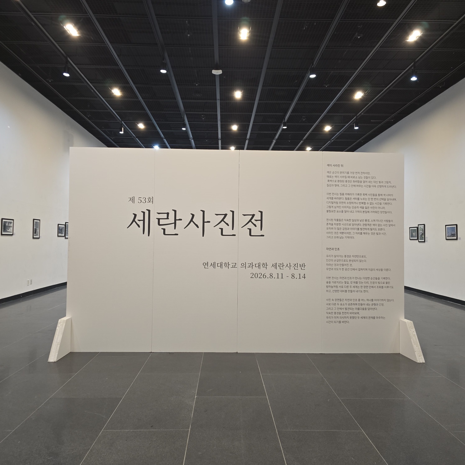 전시장 내부에 설치된 대형 백색 가벽. '제 53회 세란사진전' 제목과 전시 정보, 설명 텍스트가 인쇄되어 있음. 하단에 삼각형 지지대가 양쪽에 부착되어 자립형으로 설치됨. 뒷면으로 갤러리 공간과 액자 작품들이 보임.