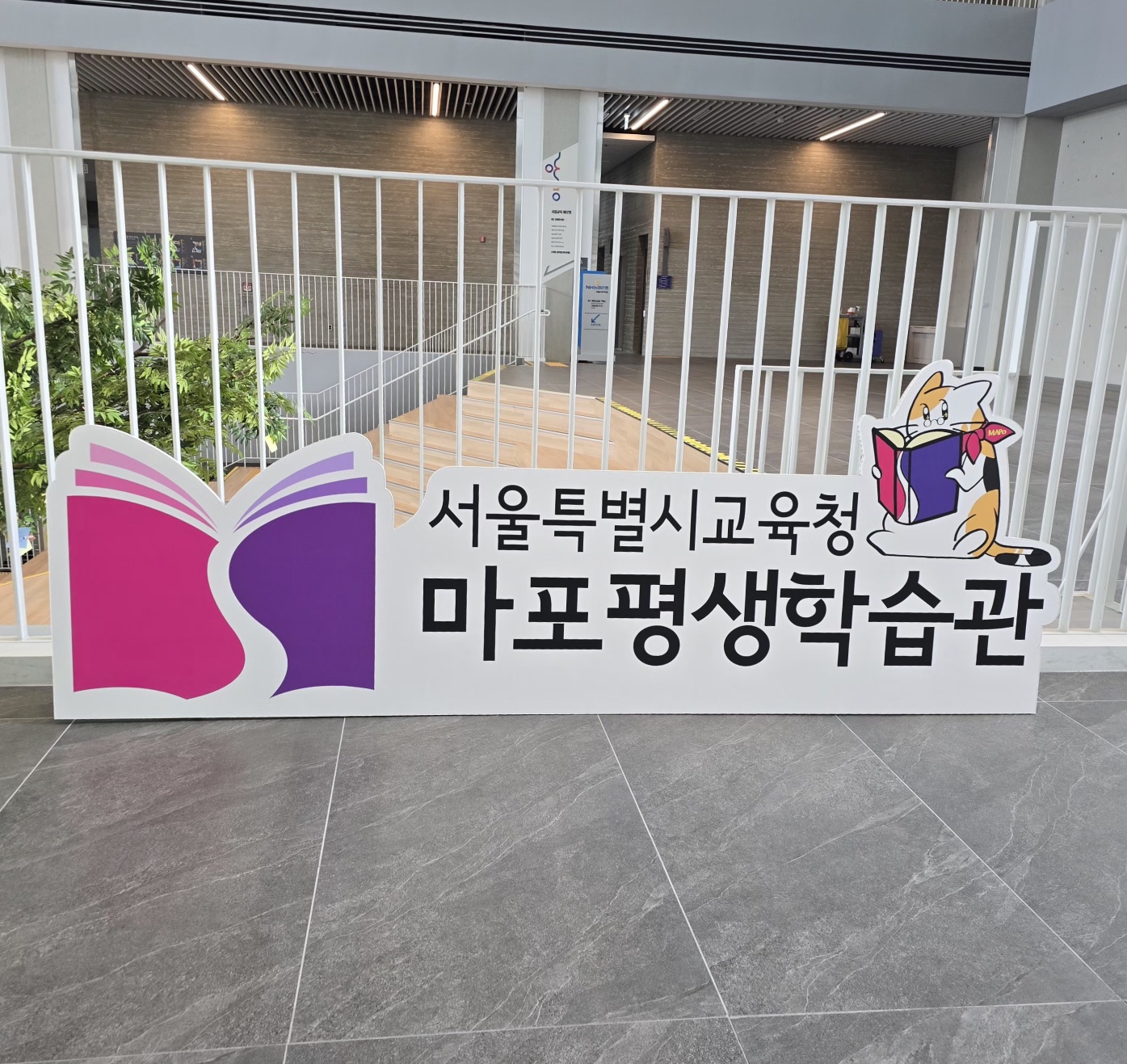 허니콤보드 평판 인쇄물. '서울특별시교육청 마포평생학습관' 문구와 책 읽는 고양이 캐릭터가 인쇄됨. 흰색 바탕에 핑크·퍼플 색상의 책 로고와 검정 글씨. 흰 펜스 앞에 설치된 모습.