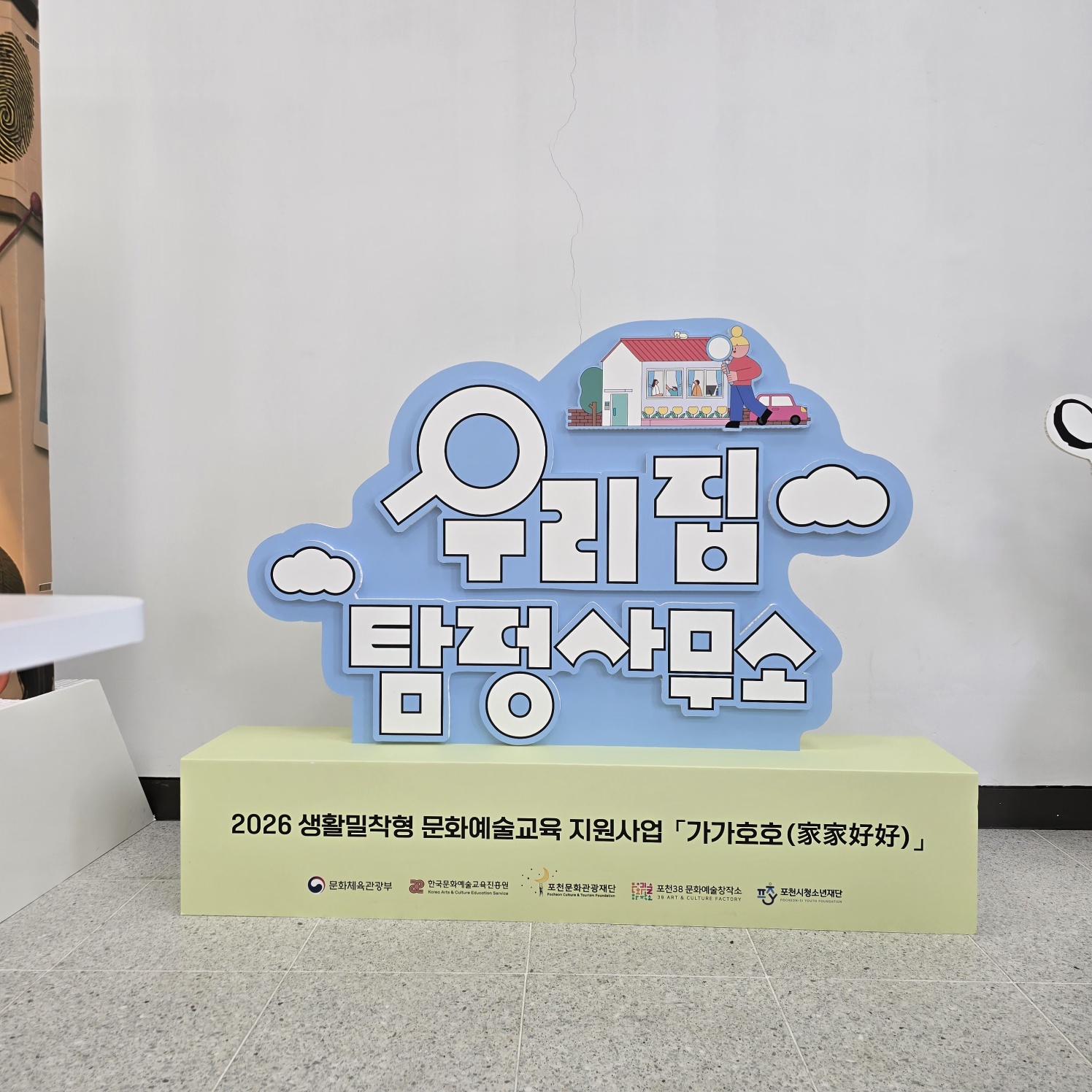 하늘색 구름 모양 배경에 '우리집 탐정사무소'라는 입체 글씨가 부착된 스카시. 상단에 집과 사람 일러스트, 돋보기 아이콘이 장식됨. 하단 연두색 박스 받침대에 '2026 생활밀착형 문화예술교육 지원사업 「가가호호(家家好好)」' 문구와 여러 기관 로고가 인쇄됨
