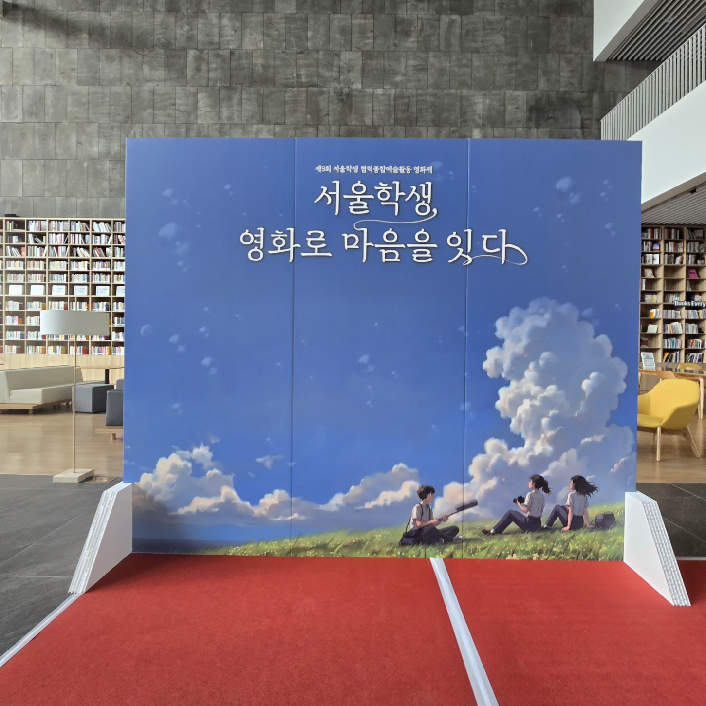 파란 하늘과 구름, 하단 초록 잔디에 학생들이 앉아 있는 일러스트가 인쇄된 배경 패널. '서울학생, 영화로 마음을 잇다' 문구가 상단에 흰색으로 새겨져 있고, 양쪽에 흰색 삼각 받침대가 보임. 뒤쪽에 도서관 책장이 보임.