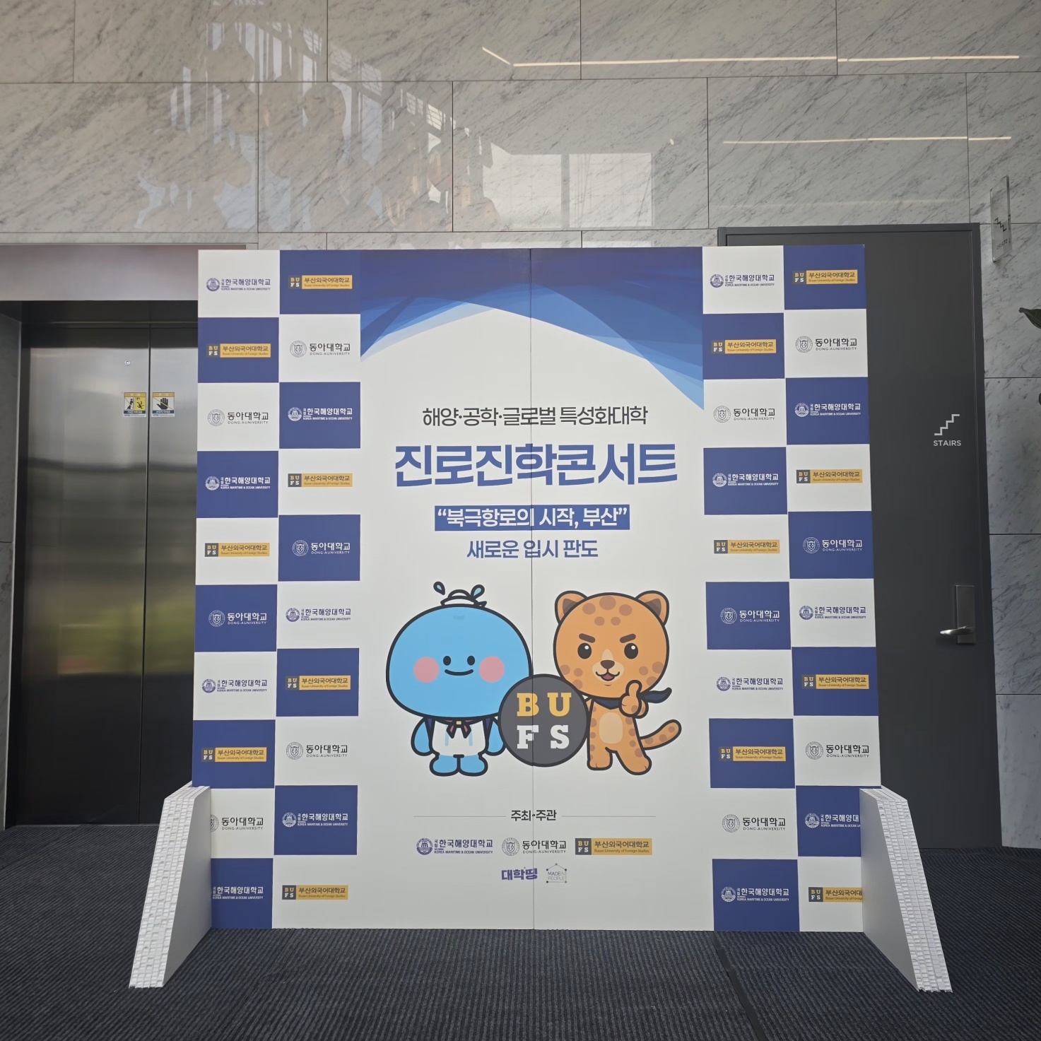체스판 형태의 청색·백색 배경에 대학 로고들이 반복 배치된 포토월. 중앙엔 '진로진학콘서트' 타이틀, 파란 곰·갈색 곰 캐릭터('BUFS'), 하단에 주최·주관·대학명 정보.