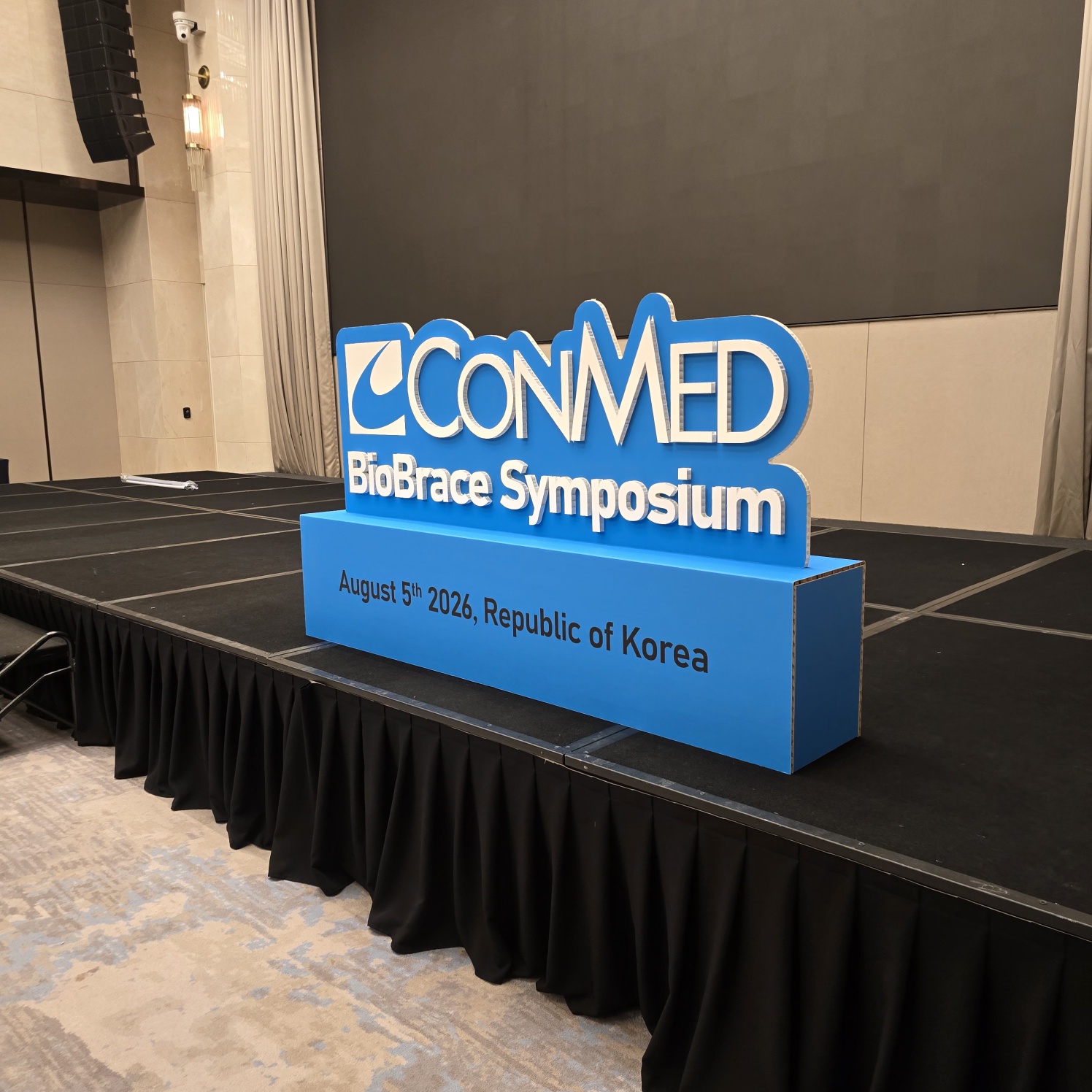 파란색 박스형 하단부와 입체 흰색 글씨로 제작된 'CONMED BioBrace Symposium' 사인물. 'August 5th 2026, Republic of Korea' 문구 포함. 검은 주름 천으로 덮인 테이블 위에 배치됨.