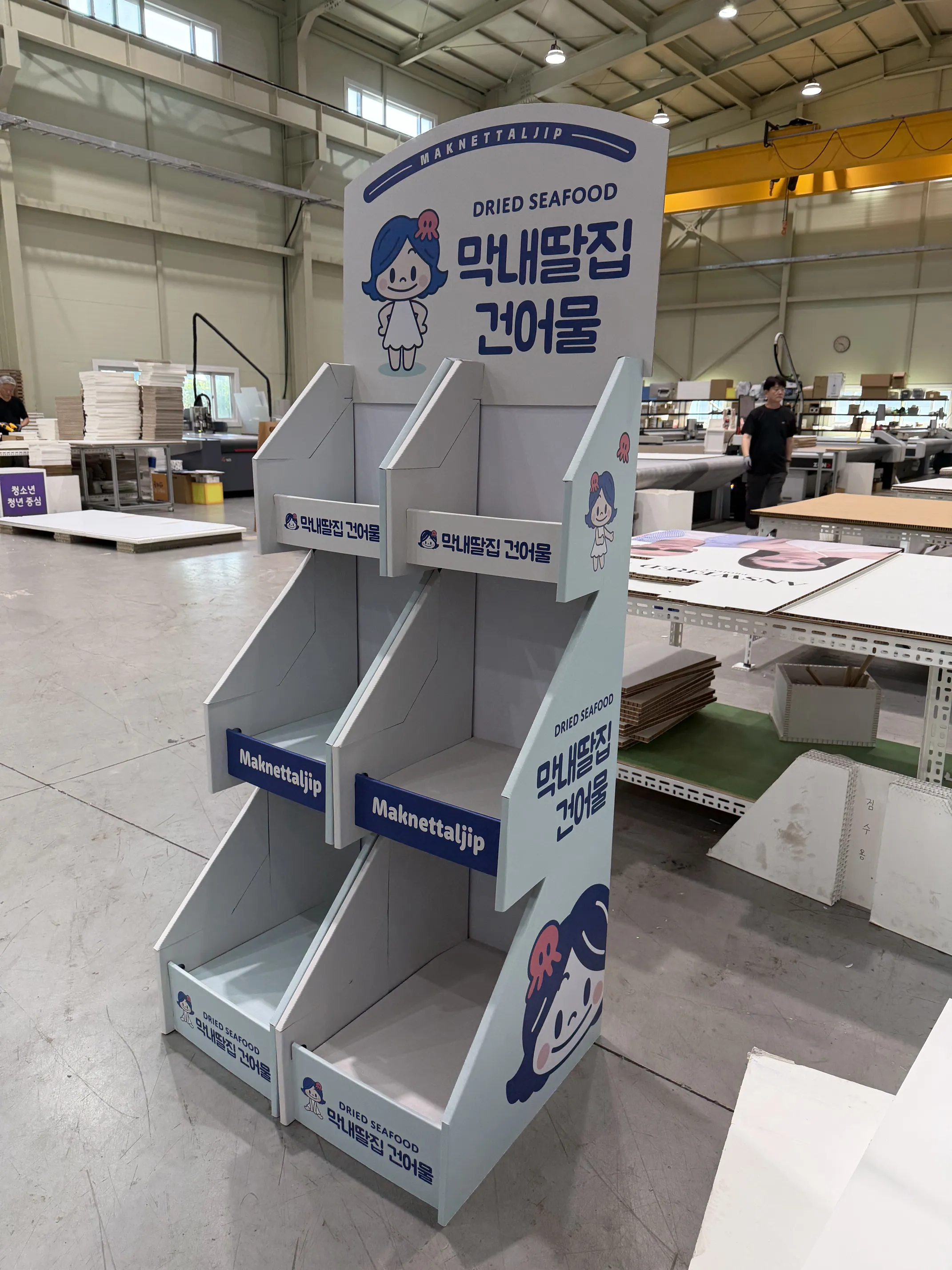 동일 매대의 측면 사진. 백색·회색 허니콤보드, 경사진 선반 3단과 바닥 1단. 측면에도 캐릭터와 'Maknettaljip' 인쇄. 십자형 구조로 4면 진열 가능.