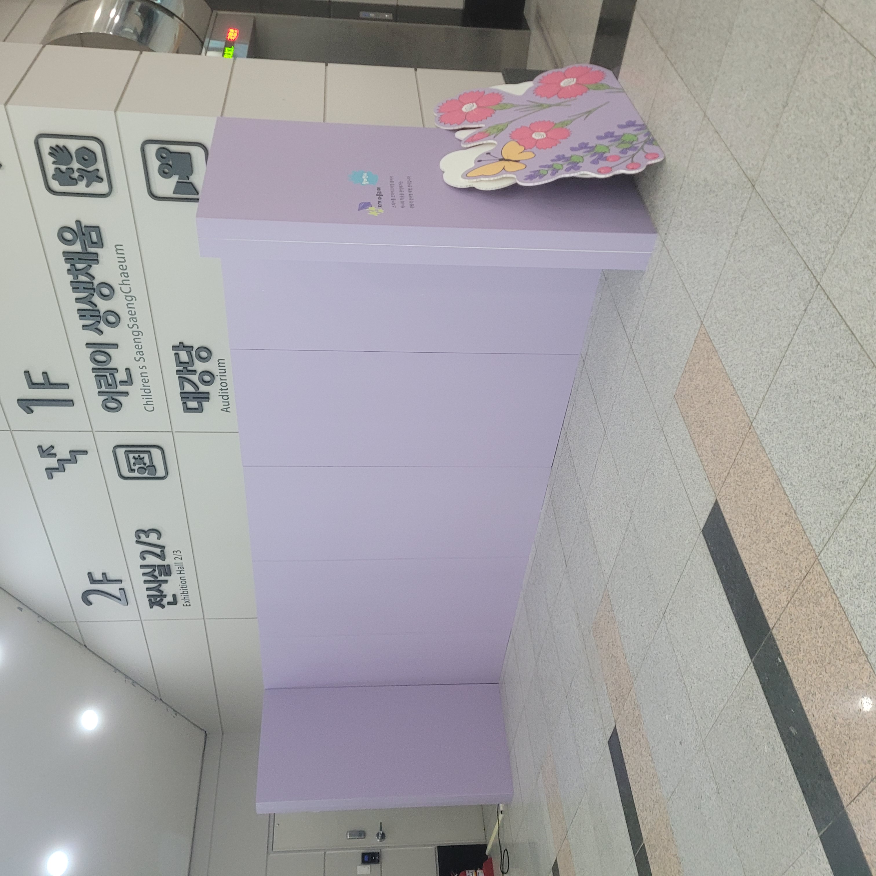 연보라색 ㄱ자형 가벽, 우측 끝에 꽃과 나비 일러스트가 인쇄된 입체 장식 부착. 가벽 상단에 로고와 텍스트 인쇄. 전시장 입구 공간 구획용.