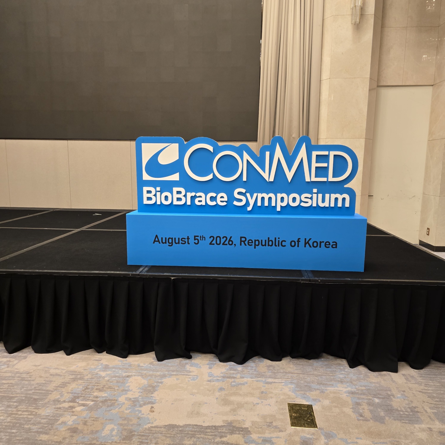 파란색 상자 위에 입체 글씨 형태로 'CONMED BioBrace Symposium'과 'August 5th 2026, Republic of Korea' 문구가 흰색으로 인쇄된 행사용 사인물. 검은 천으로 덮인 테이블 위에 설치됨.