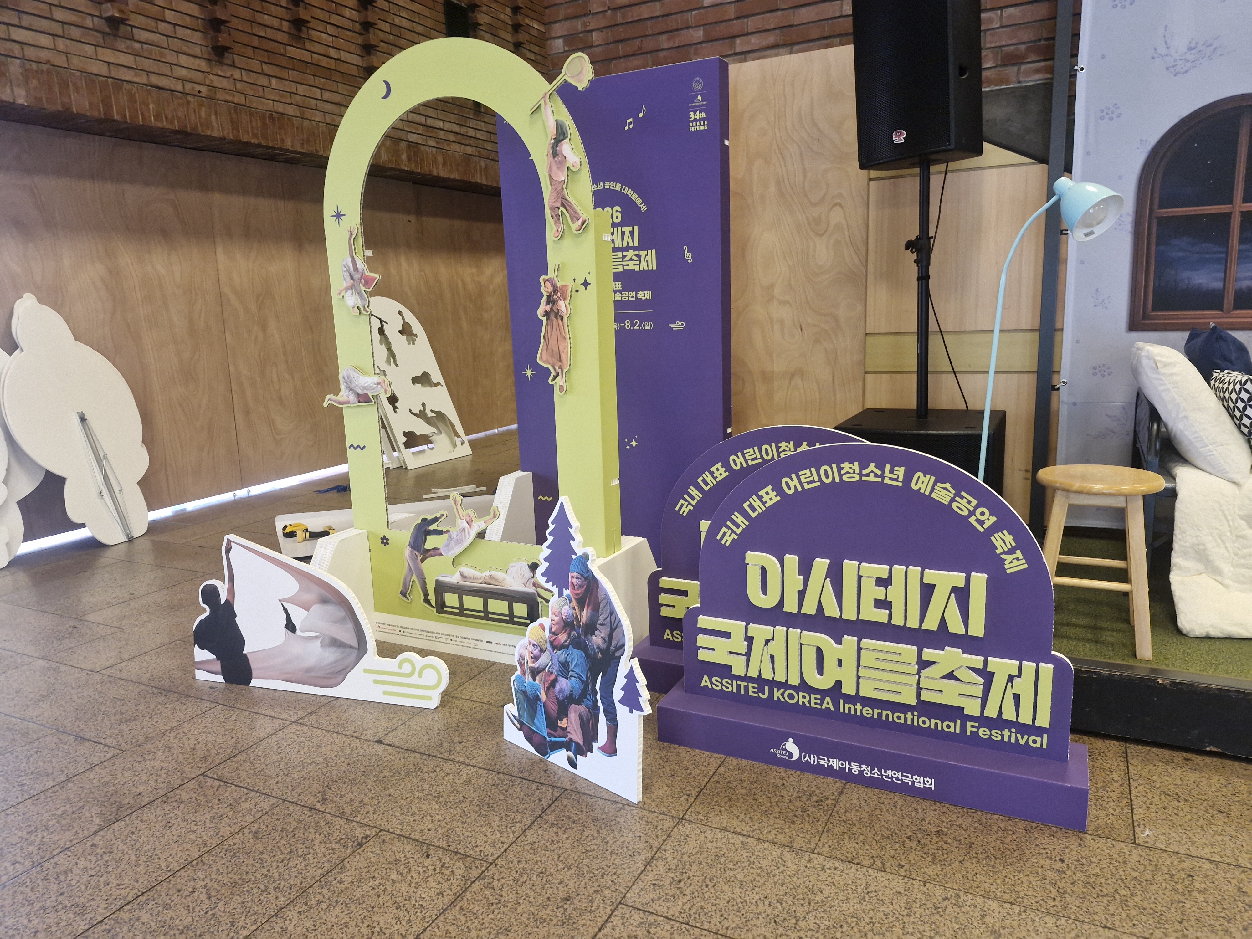 달과 아치형 게이트 모양의 노란색·보라색 입체 포토존 구조물, 'ASSITEJ KOREA International Festival' 문구 인쇄, 하단에 인물 등신대 패널 여러 개 배치