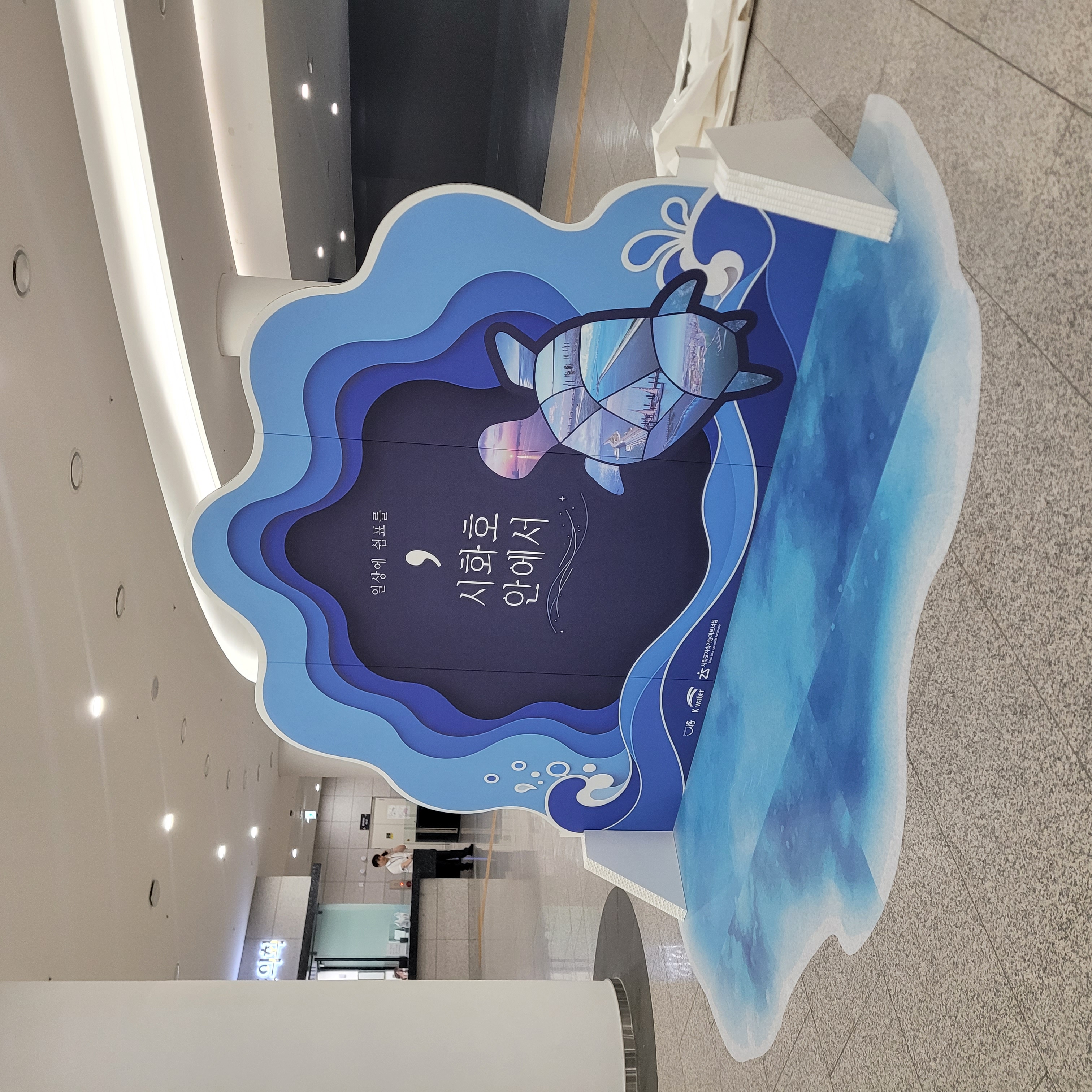 사진0과 동일한 포토존을 측면에서 촬영. 청색 물결 레이어 구조와 거북이 일러스트, '시화호 안에서' 문구 확인. 하단 물결 바닥 그래픽과 화이트 테두리 마감 처리.