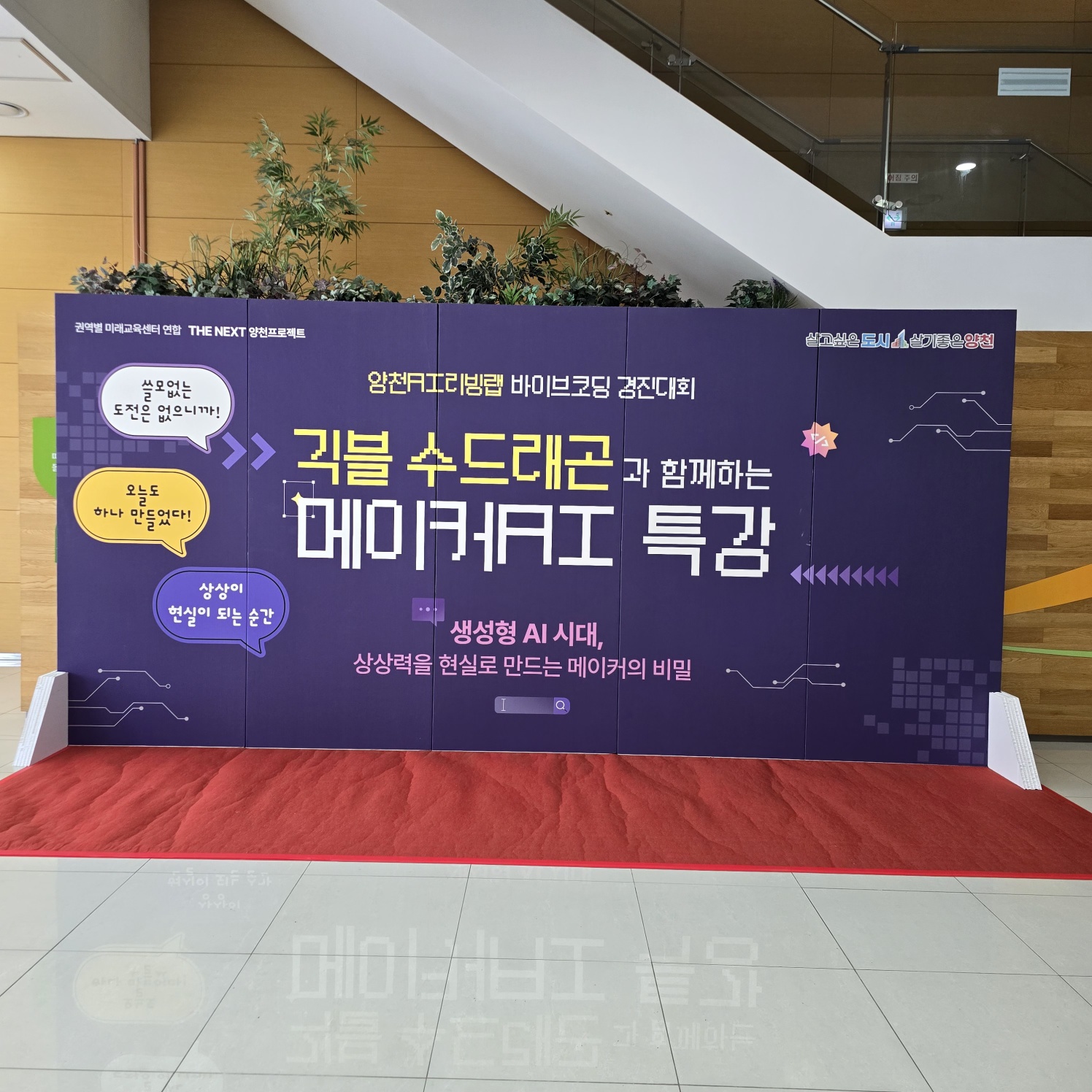 보라색 바탕에 노란색·흰색 글씨로 '귀블 수드레곤과 함께하는 메이커AI 특강' 등이 인쇄된 대형 가벽. 상단에 화분이 놓여 있고, 앞쪽에 빨간 카펫이 깔려 있음.