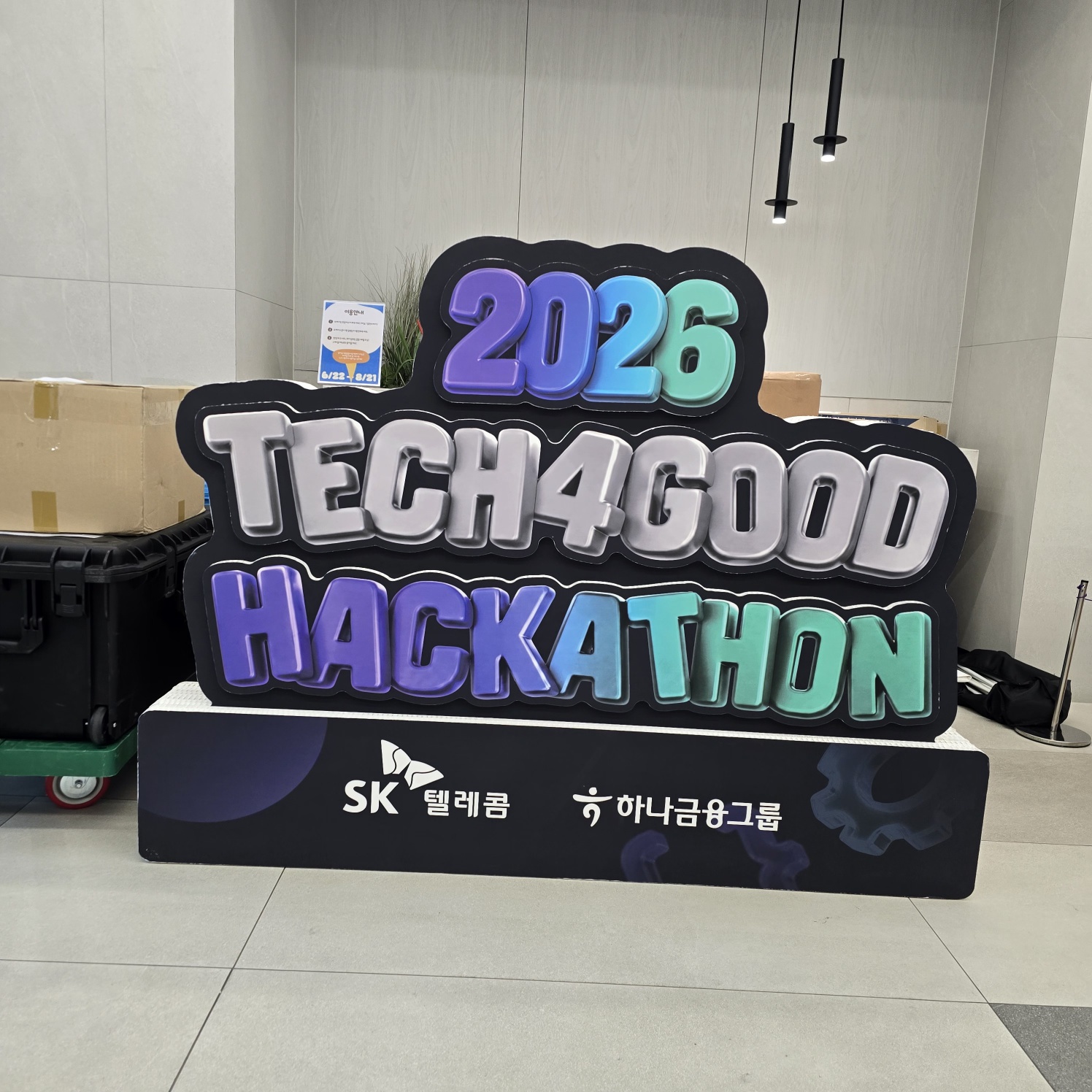 검은색 테두리 로고형 입체 조형물. '2026 TECH4GOOD HACKATHON' 텍스트가 그래디언트 색상(보라-파랑-초록)으로 인쇄되어 있으며, 하단에 'SK 텔레콤', '하나금융그룹' 로고가 인쇄된 검정 받침대가 있는 포토존 설치물