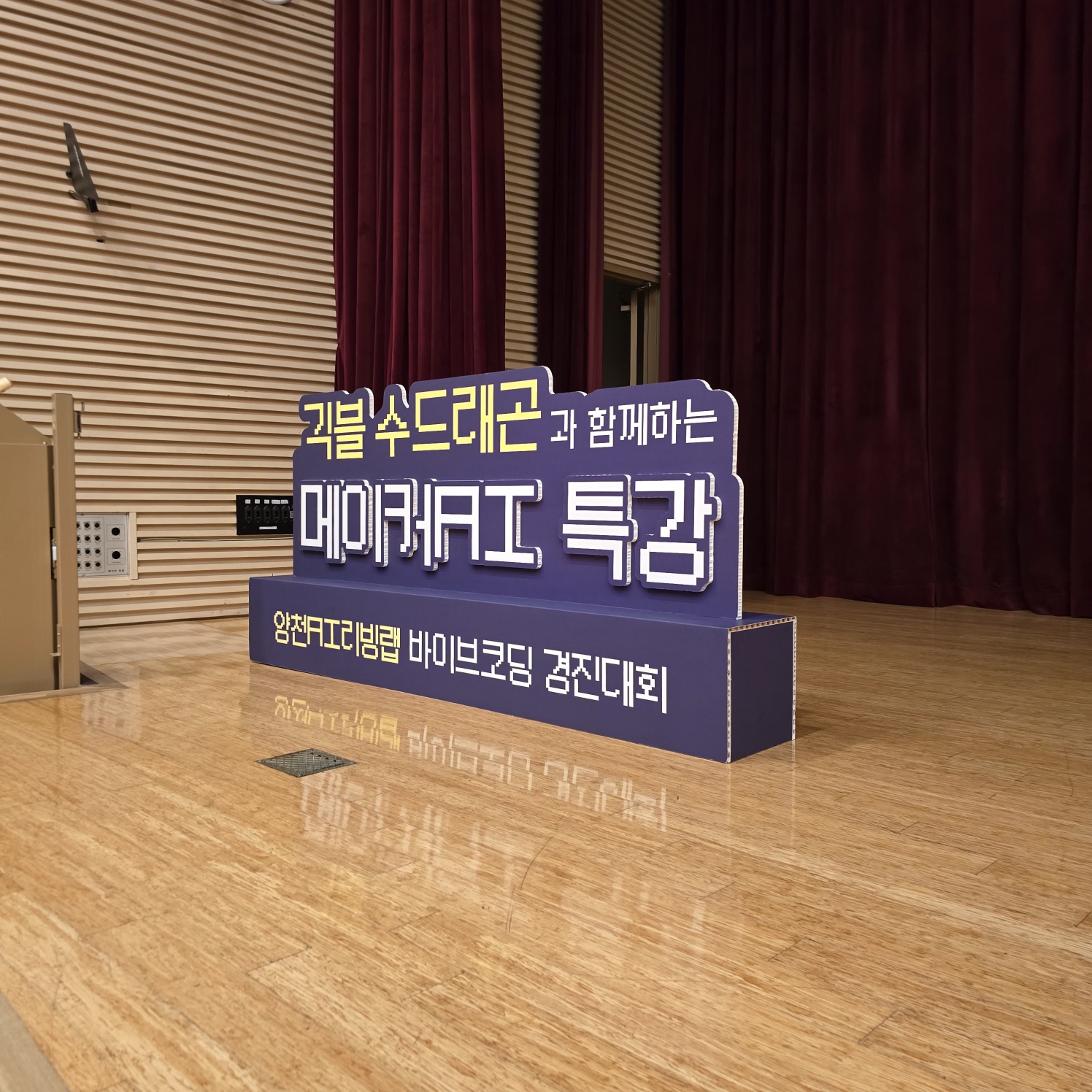 보라색 바탕에 '귀블 수드레곤과 함께하는 메이커AI 특강' 문구가 입체로 제작된 스카시. 아래 긴 받침 박스에 '양천AI디지털랩 바이브코딩 경진대회' 인쇄. 강당 바닥에 설치.