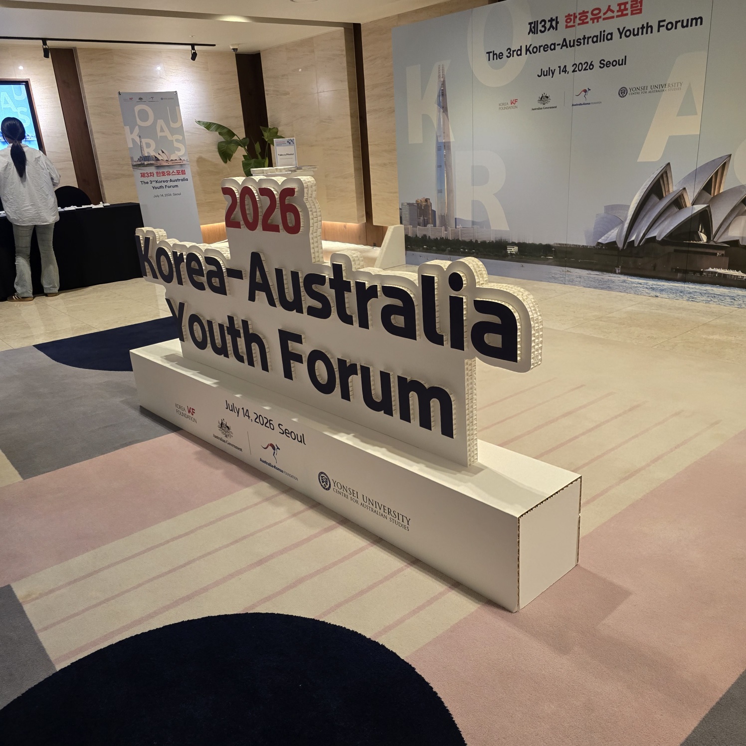 흰색 아크릴 받침 위에 진한 파란색 입체 글씨로 'Korea-Australia Youth Forum' 텍스트가 있으며, '2026' 숫자가 분홍색으로 표현되어 있음. 하단부에 'July 14, 2026 Seoul'과 YONSEI UNIVERSITY 등 로고가 인쇄된 흰색 받침판