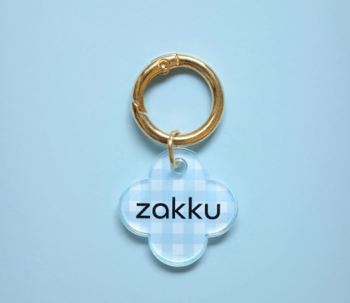 하늘색 체크무늬 패턴의 아크릴 네잎클로버 형태 키링. 검은색으로 'zakku' 문구가 인쇄되어 있고, 금색 키링 고리에 연결됨