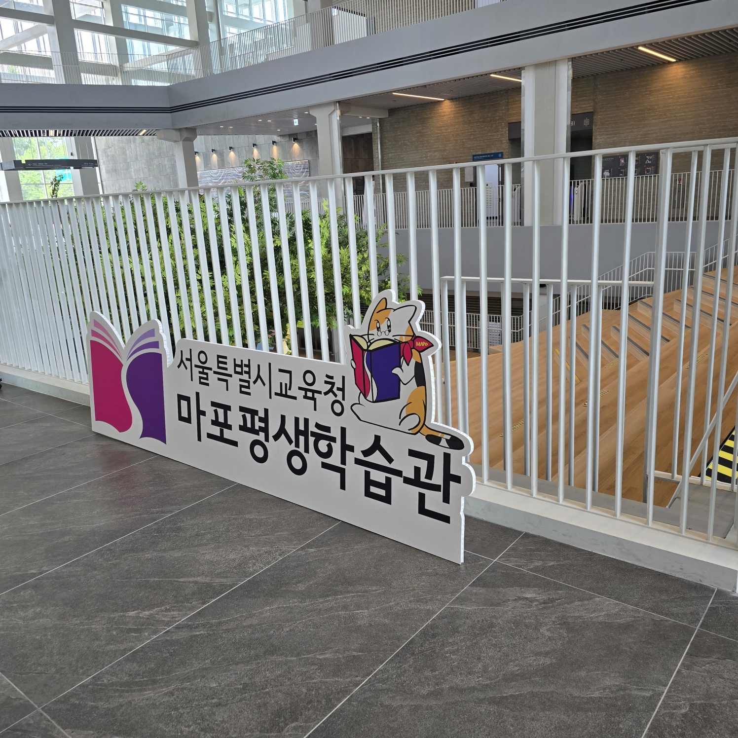 허니콤보드 평판 인쇄물의 측면 각도. 흰색 펜스 앞에 거치되어 있으며, 앞면에 '서울특별시교육청 마포평생학습관' 로고와 책 캐릭터가 선명하게 인쇄됨. 단면 인쇄로 보임.