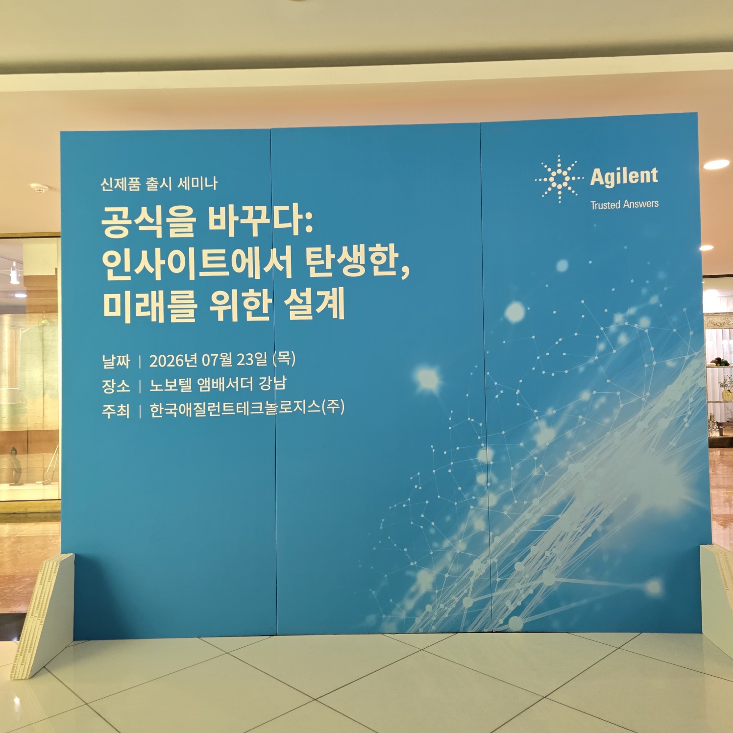 파란색 바탕에 'Agilent' 로고와 세미나 정보가 인쇄된 대형 패널. '공식을 바꾸다: 인사이트에서 탄생한, 미래를 위한 설계' 문구, 날짜·장소 표기. 하단에 흰색 허니콤 구조 지지대가 보이며 실내 세미나 입구에 설치됨.