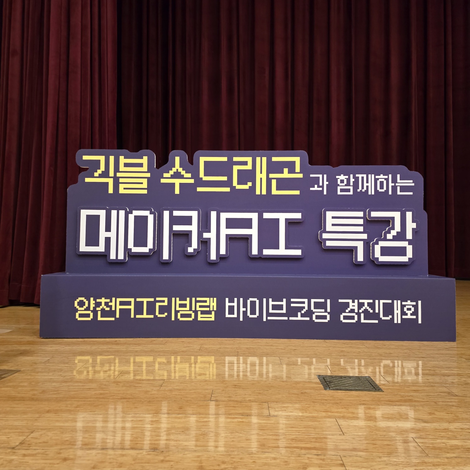 보라색 바탕에 노란색·흰색으로 '귀블 수드레곤과 함께하는 메이커AI 특강' 문구가 입체적으로 제작된 스카시. 하단 받침 박스에 작은 글씨가 추가되어 무대 위에 자립 설치됨.