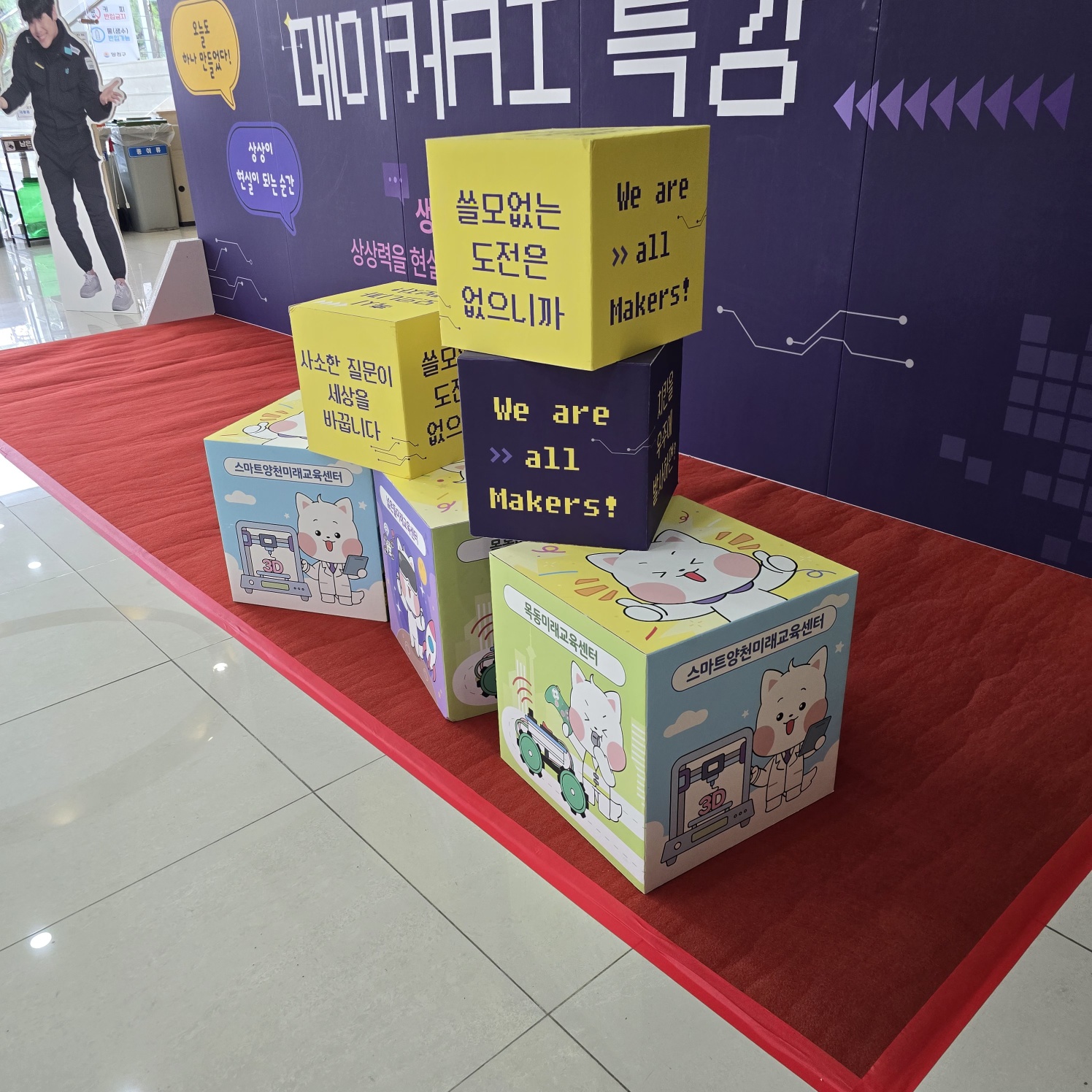 노란색·보라색 큐브에 '쓸모없는 도전은 없으니까', 'We are all Makers!' 등 문구와 캐릭터가 인쇄됨. 총 6개가 쌓여 빨간 카펫 위 전시됨.