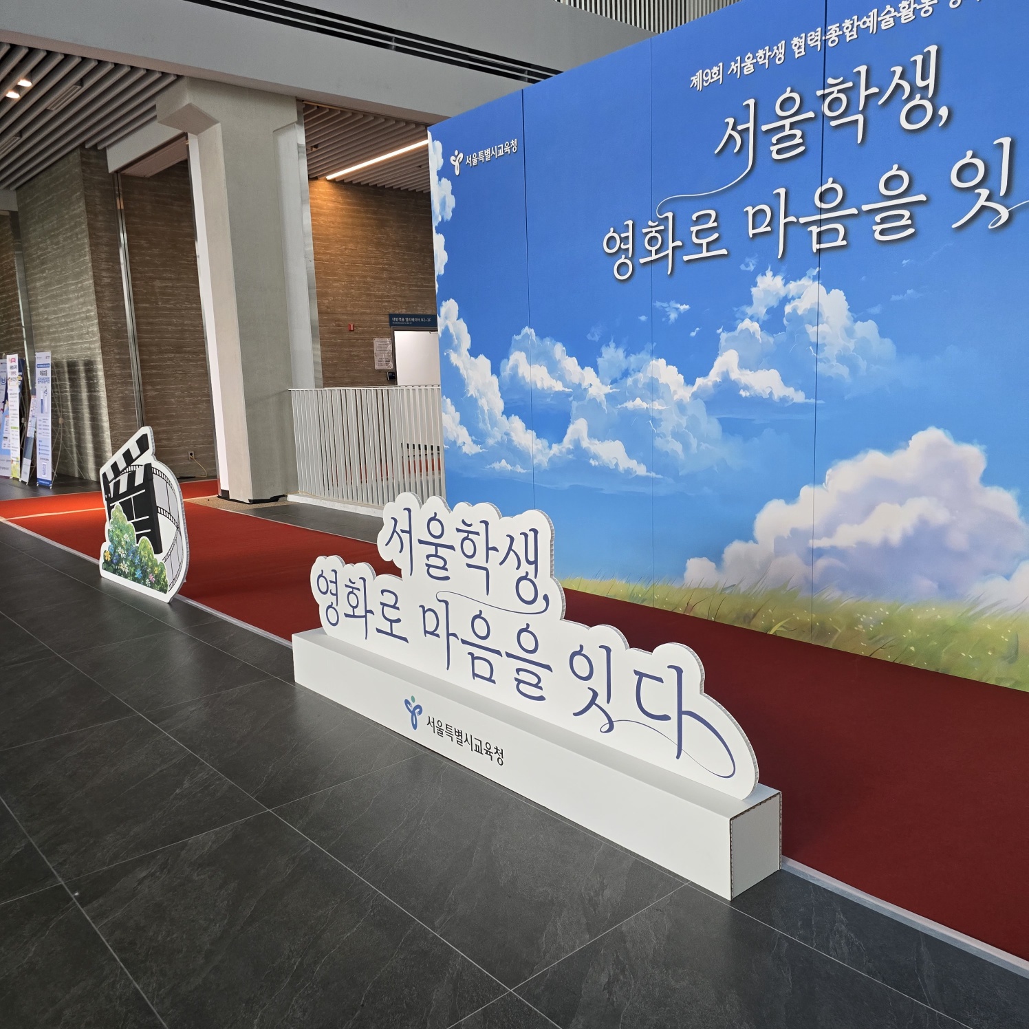 같은 파란 하늘 배경 패널과 구름 모양 간판, 왼쪽에 영화 필름릴 장식물이 배치된 모습. 빨간 카펫이 깔려 있고, 배경에 실내 복도와 난간이 보임.