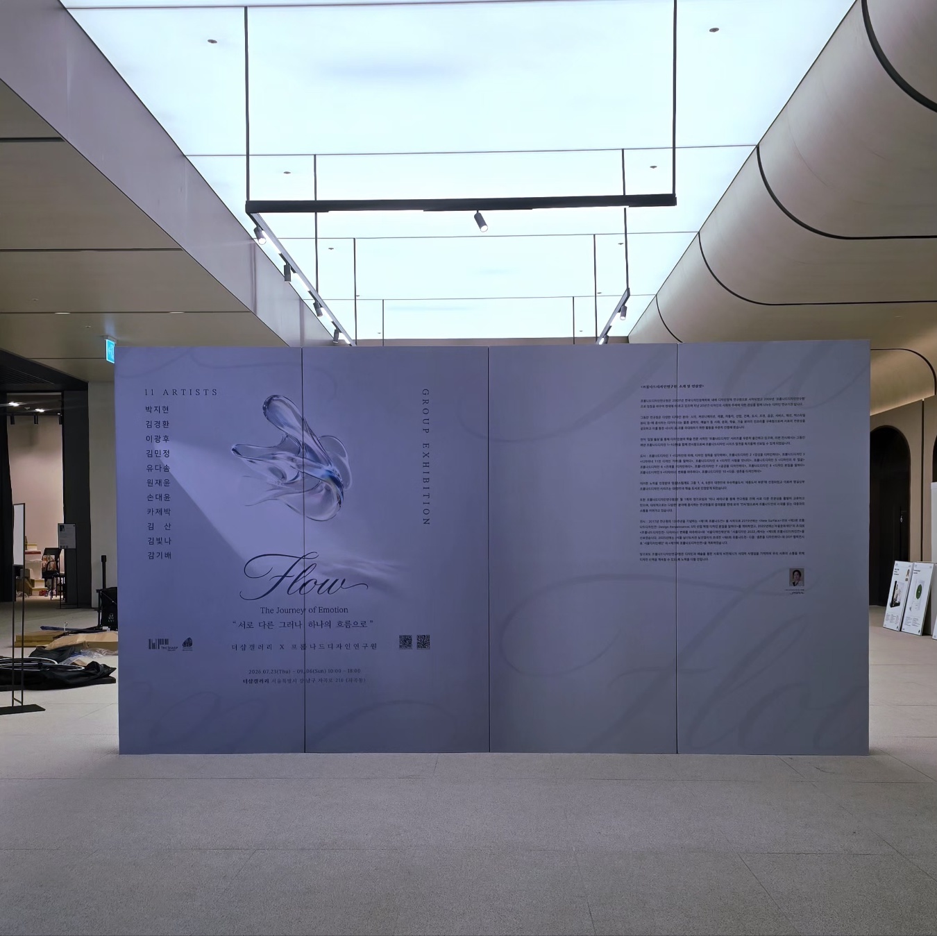 연보라색 대형 평면 가벽 3장 연결, 좌측에 '11 ARTISTS' 및 작가명 세로 나열, 중앙에 투명한 손 이미지와 'Flow' 캘리그래피 인쇄, 우측에 전시 소개 텍스트 배치