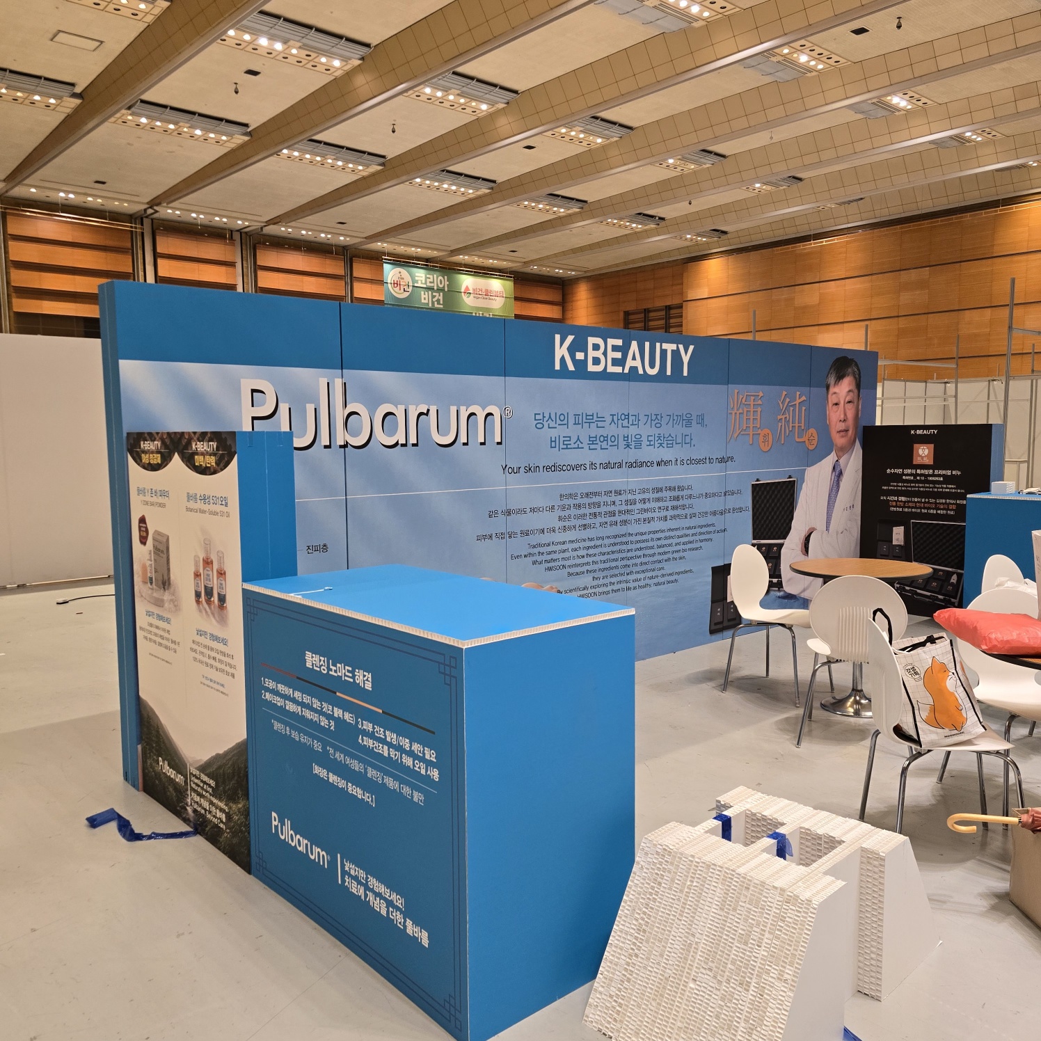 정면 근접 촬영. 청색 허니콤보드 카운터, 상단 백색 테이블, 'K-BEAUTY', 'Pulbarum' 로고와 모델 사진·제품 이미지 대형 인쇄, 내부에 흰색 의자와 테이블 배치, 바닥에 컬러 쿠션 보임