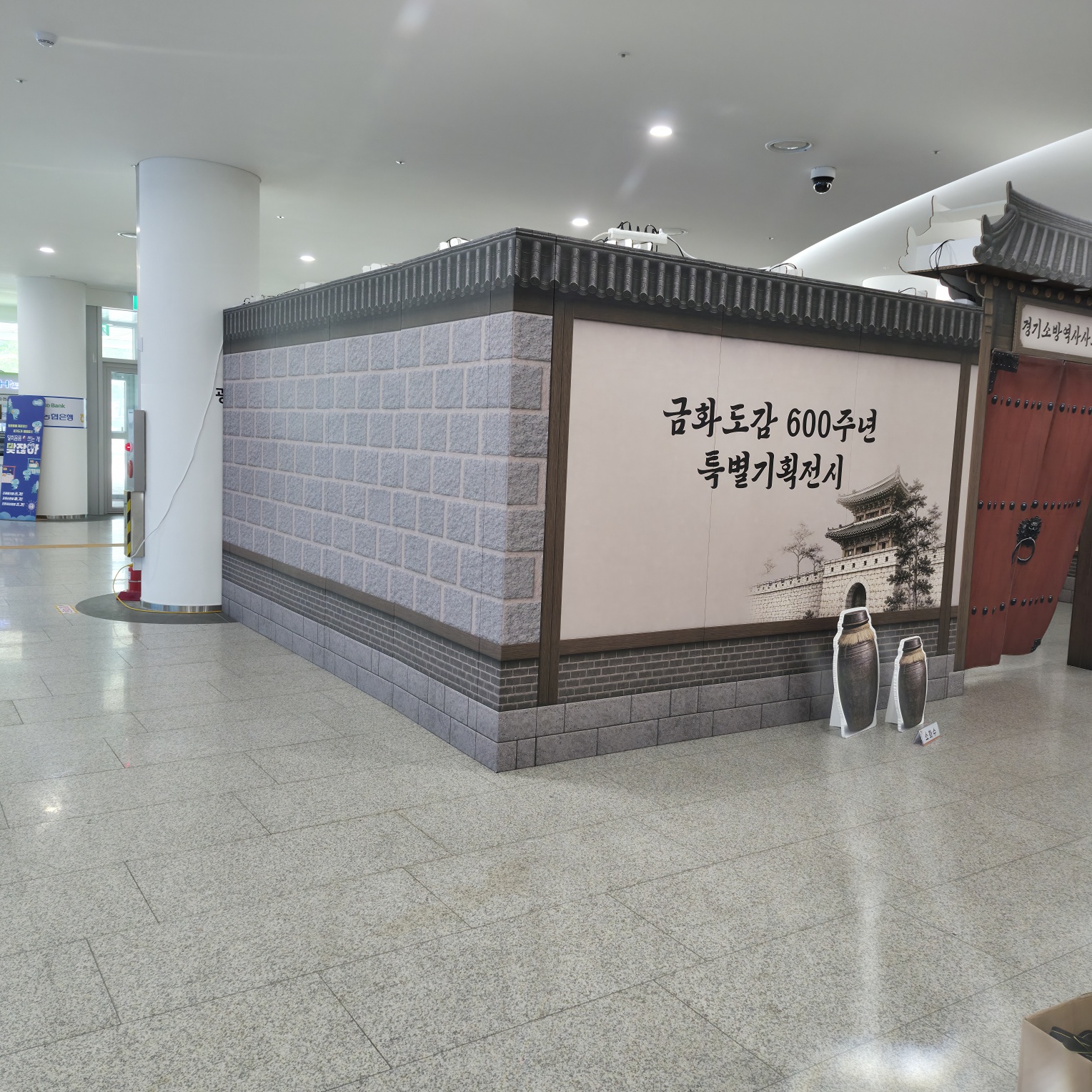 한옥 전통 건축 형태의 포토존 구조물. 목재 프레임과 석재 질감의 측면, 검은색 기와지붕 장식이 있으며, 한쪽 면에는 흰색 배경에 한글 텍스트와 건축물 이미지 인쇄, 다른 쪽은 붉은색 목재 문짝 형태임