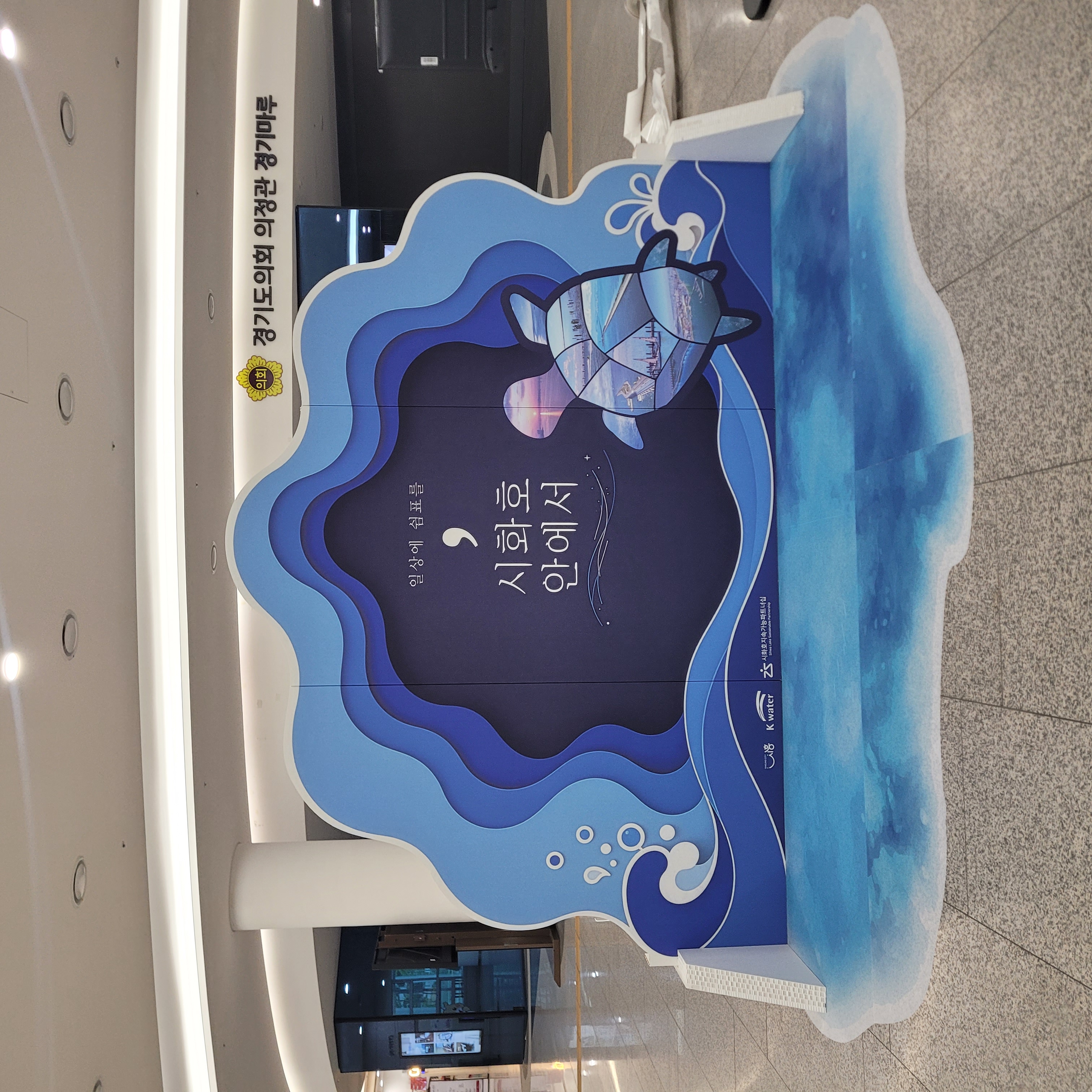 파도와 물결 형태로 레이어 구조를 이룬 청색 계열 포토존. 중앙에 '일상에 쉼표를, 시화호 안에서' 문구와 거북이·풍경 일러스트 인쇄. 하단 물결 바닥 그래픽 포함. 의정관 경기마루 명칭 표기.