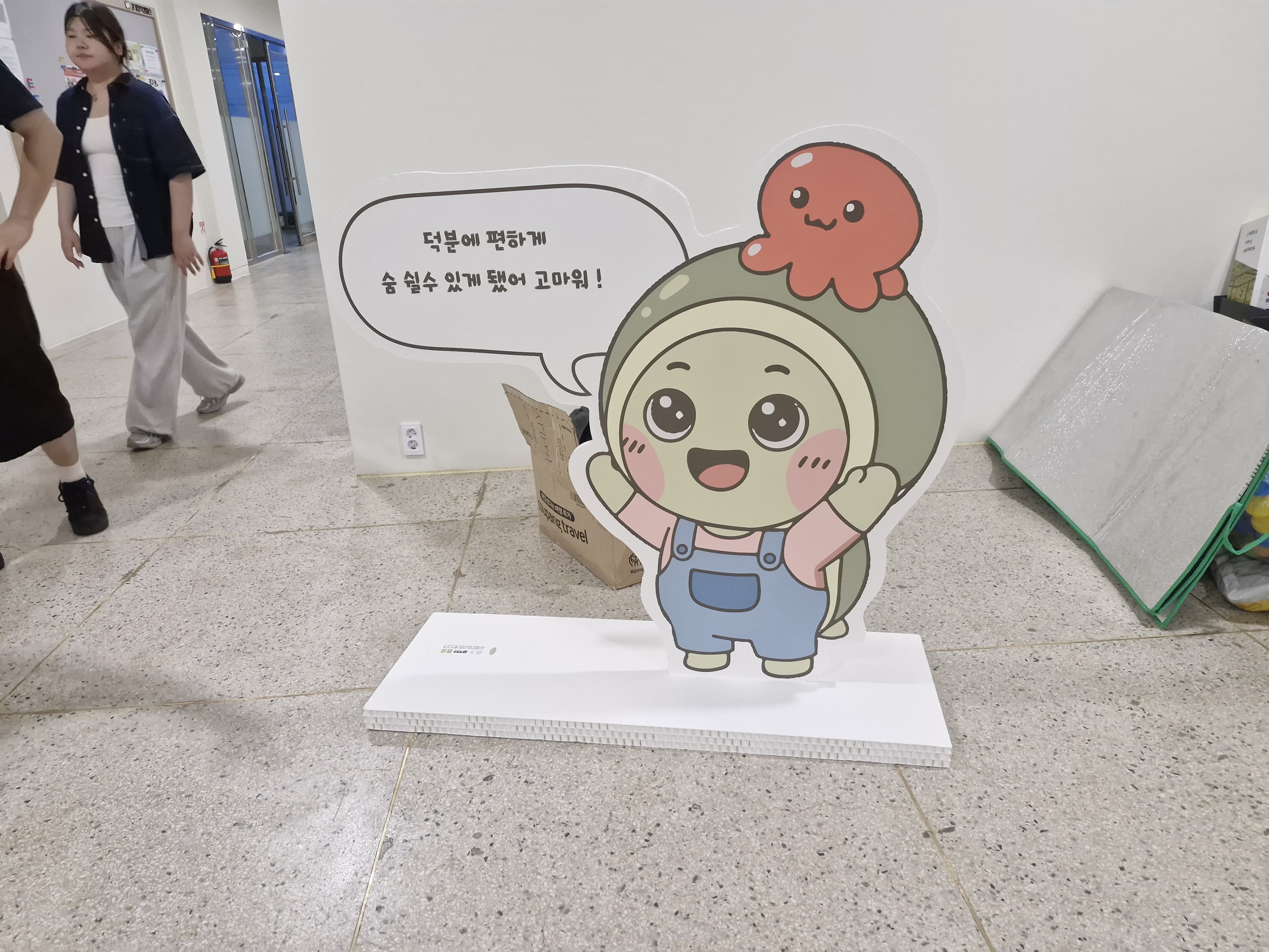 가방 메고 웃는 어린이 캐릭터 스카시. 머리 위에 문어 캐릭터가 얹혀 있고, 말풍선에 '덕분에 평강'게 술 실속 있'게 댔어 고마워!' 문구. 허니콤보드 받침대에 자립.