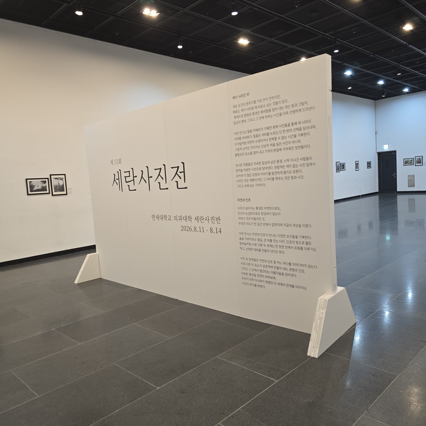 사진 0과 동일한 가벽의 측면 각도. '세란사진전' 타이틀과 전시 정보 텍스트가 인쇄된 백색 가벽. 하단 삼각 지지대 구조가 명확히 보이며, 뒤편으로 전시 공간이 이어짐.