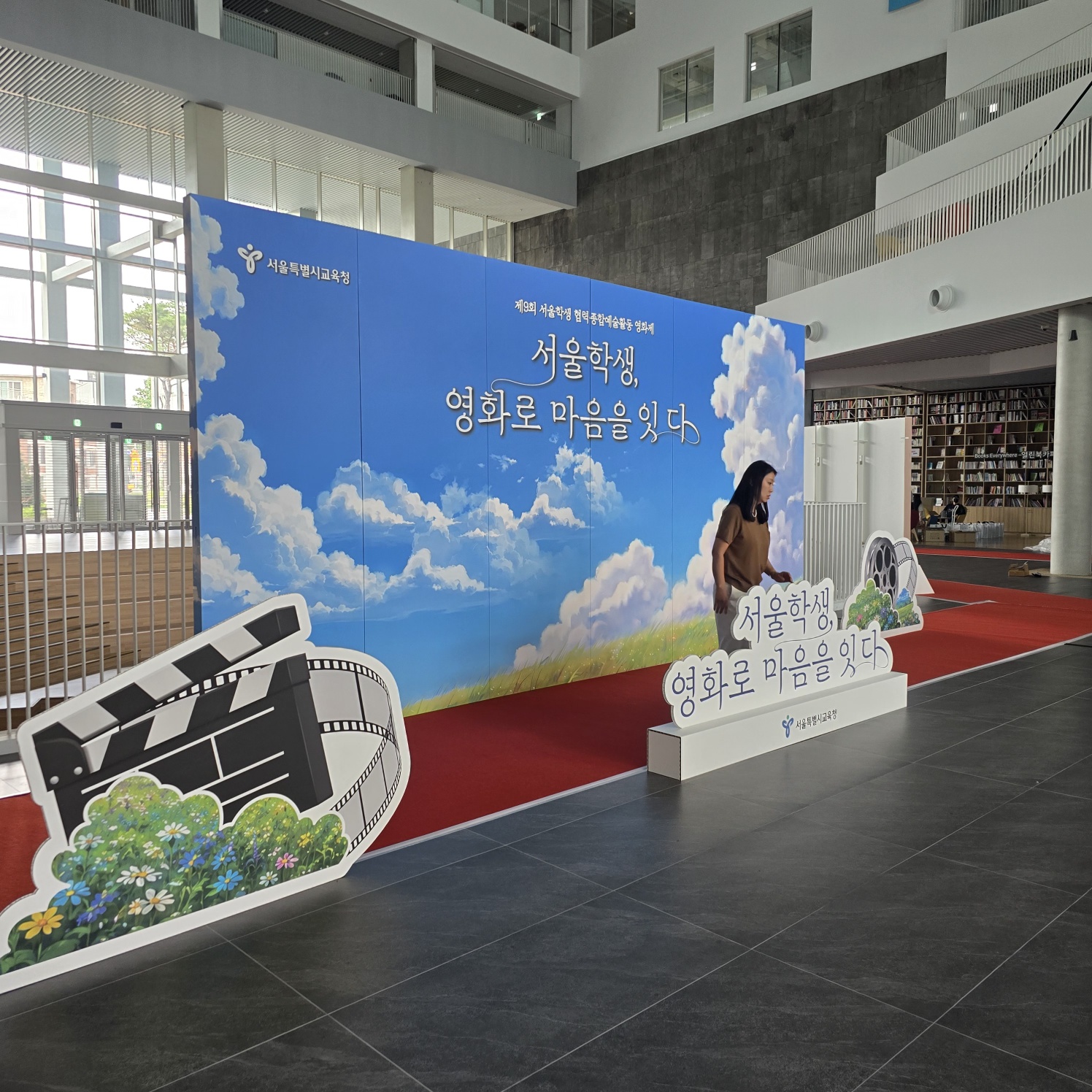 같은 파란 하늘 배경 패널 앞에 영화 필름릴과 클래퍼보드 모양의 입체 장식물이 왼쪽에, '서울학생, 영화로 마음을 잇다' 문구가 새겨진 구름 모양 흰색 스탠드형 소품이 오른쪽에 설치됨. 사람이 포토존 앞에 서 있음.