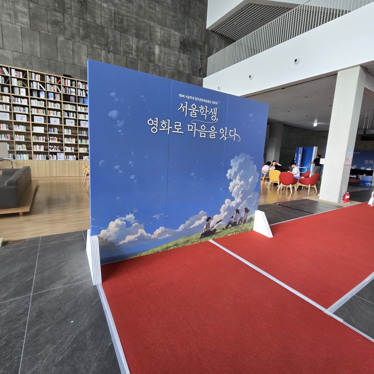 같은 배경 패널의 뒷면 또는 측면 각도. 잔디밭에 학생들이 앉아 있는 일러스트가 보이고, 양쪽에 흰색 삼각 받침대가 설치됨. 뒤쪽에 도서관 공간과 책상, 의자가 보임.