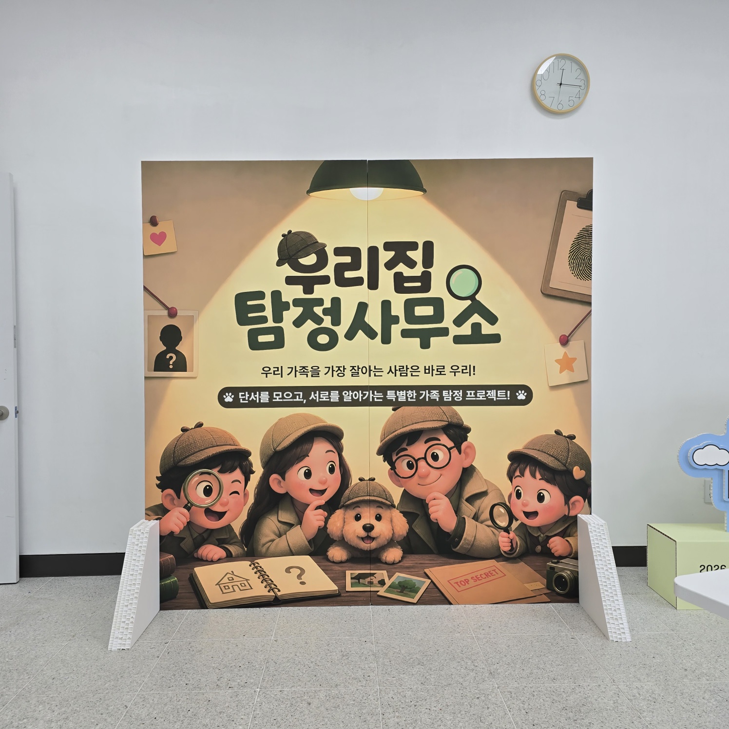 동화책 펼친 형태의 자립형 포토존. '우리집 탐정사무소'라는 큰 타이틀과 함께 탐정 모자를 쓴 어린이 캐릭터 5명과 강아지 일러스트가 인쇄됨. 조명 효과, 액자, 카메라 등 탐정 테마 소품들이 배경에 그려져 있음. 하단에 '단서를 모으고, 서로를 알아가는 특별한 가족 탐정 프로젝트' 문구. 좌우 흰색 허니콤 구조 지지대로 세워짐